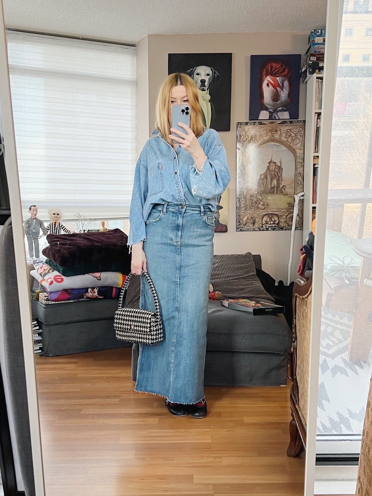 A blonde woman is wearing a denim maxi skirt, denim shirt, ballet flats, and a houndstooth bag.