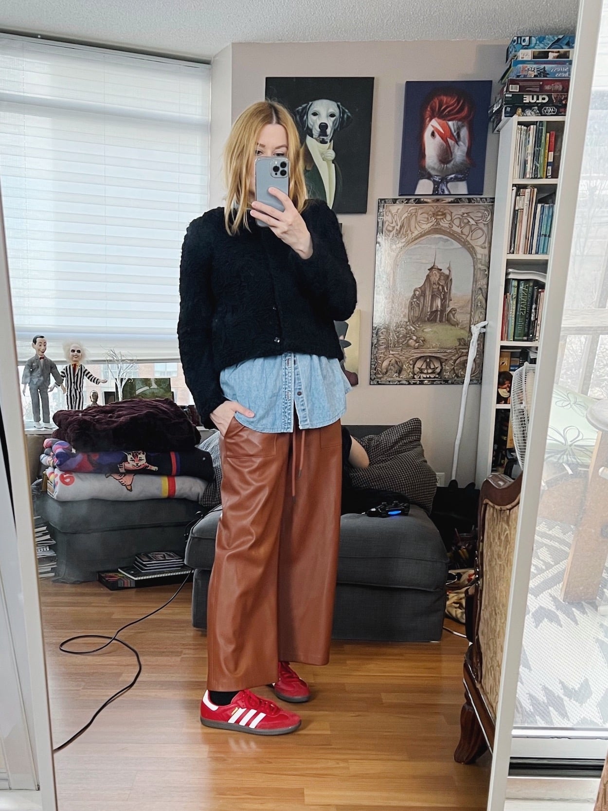 A blonde woman is wearing brown leather pants, a denim shirt layered under a black cropped jacket, and red Adidas.