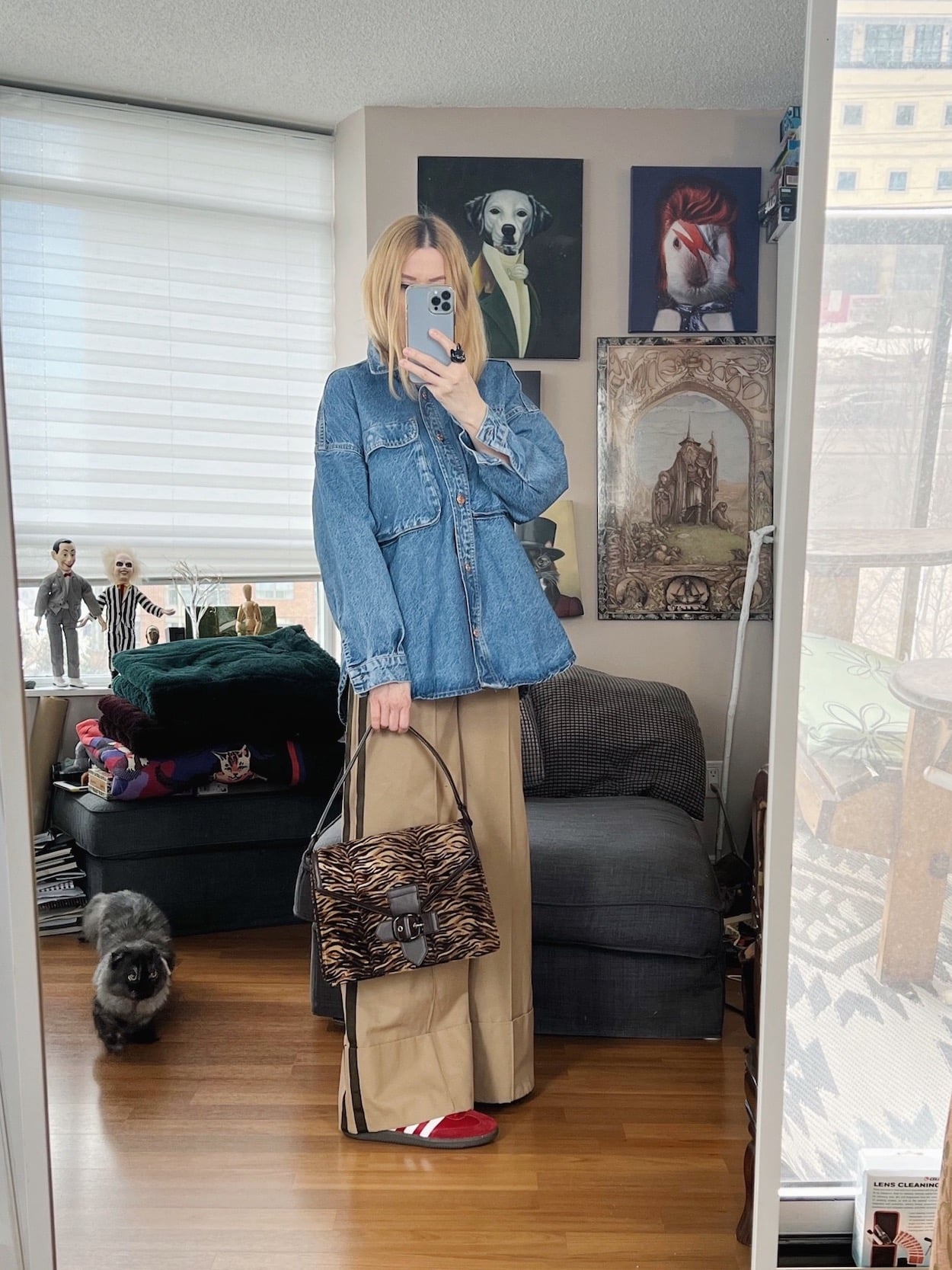 A blonde woman is wearing a denim shirt, wide leg trousers, red Adidas Sambas, and a vintage animal print bag.