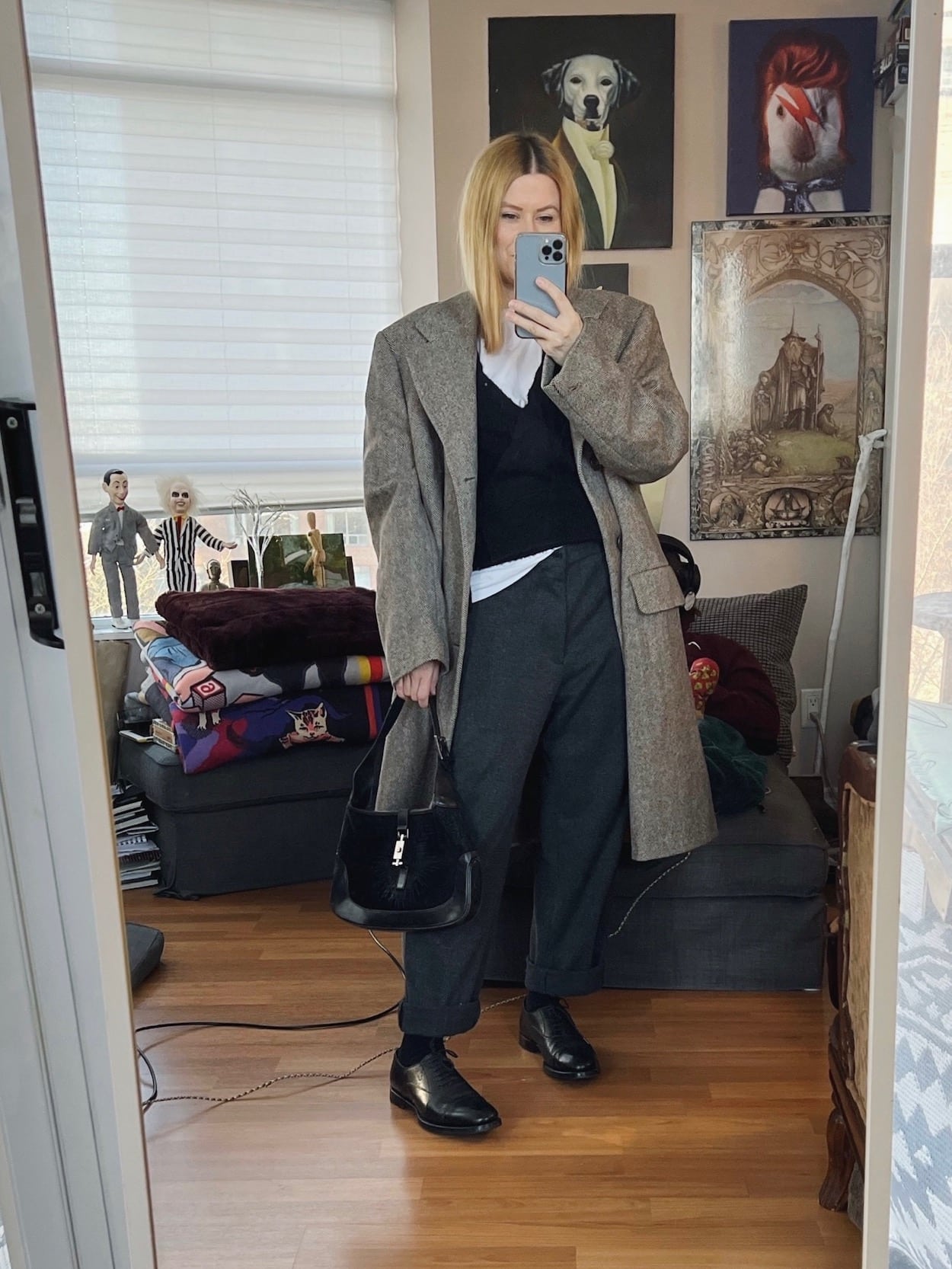 A blonde woman is wearing a white t-shirt, black cardigan, men's trousers, oxfords, a vintage men's coat, and a vintage Gucci bag.