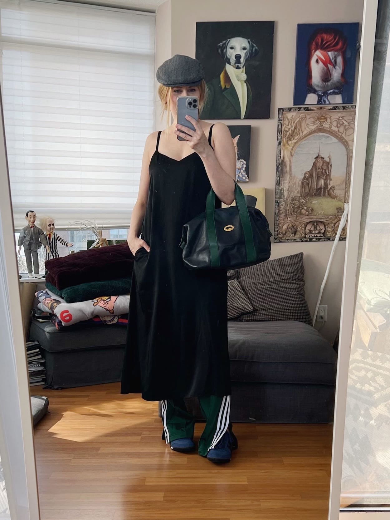 A blonde woman is wearing a black slip dress over green Adidas track pants, a driving cap, blue Onitsuka Tigers, and a vintage Longchamp bag.