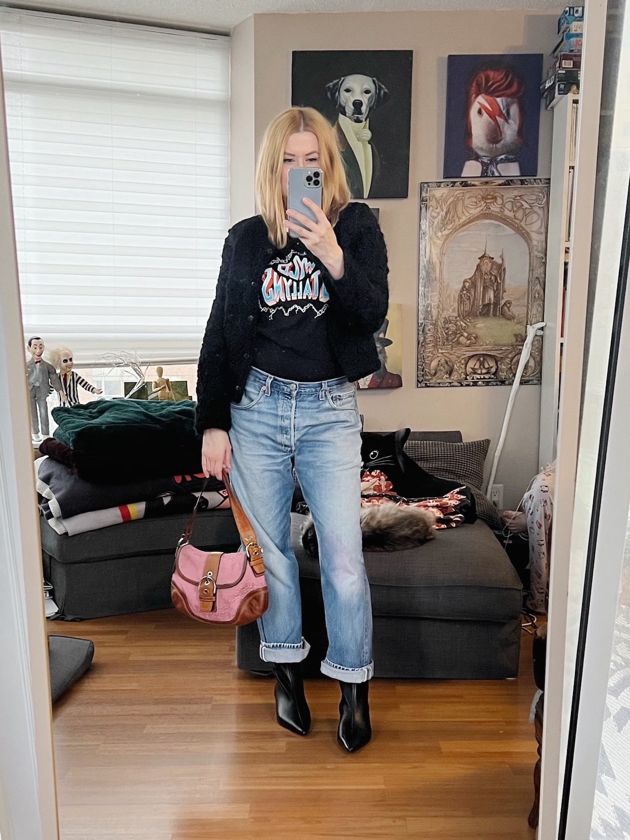 A blonde woman is wearing a wyld stallyns shirt, black cropped jacket, vintage Levi's, kitten heel boots, and a vintage coach bag.