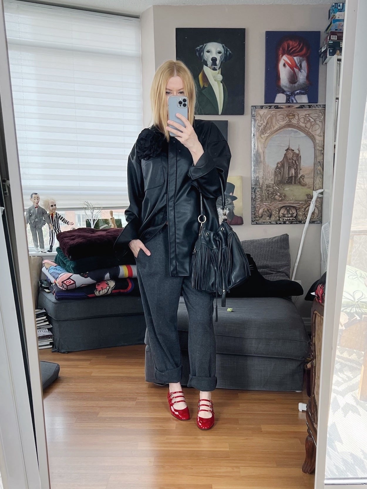 A blonde woman is wearing a faux leather shirt, grey trousers, red Mary Janes, and a fringe bag.