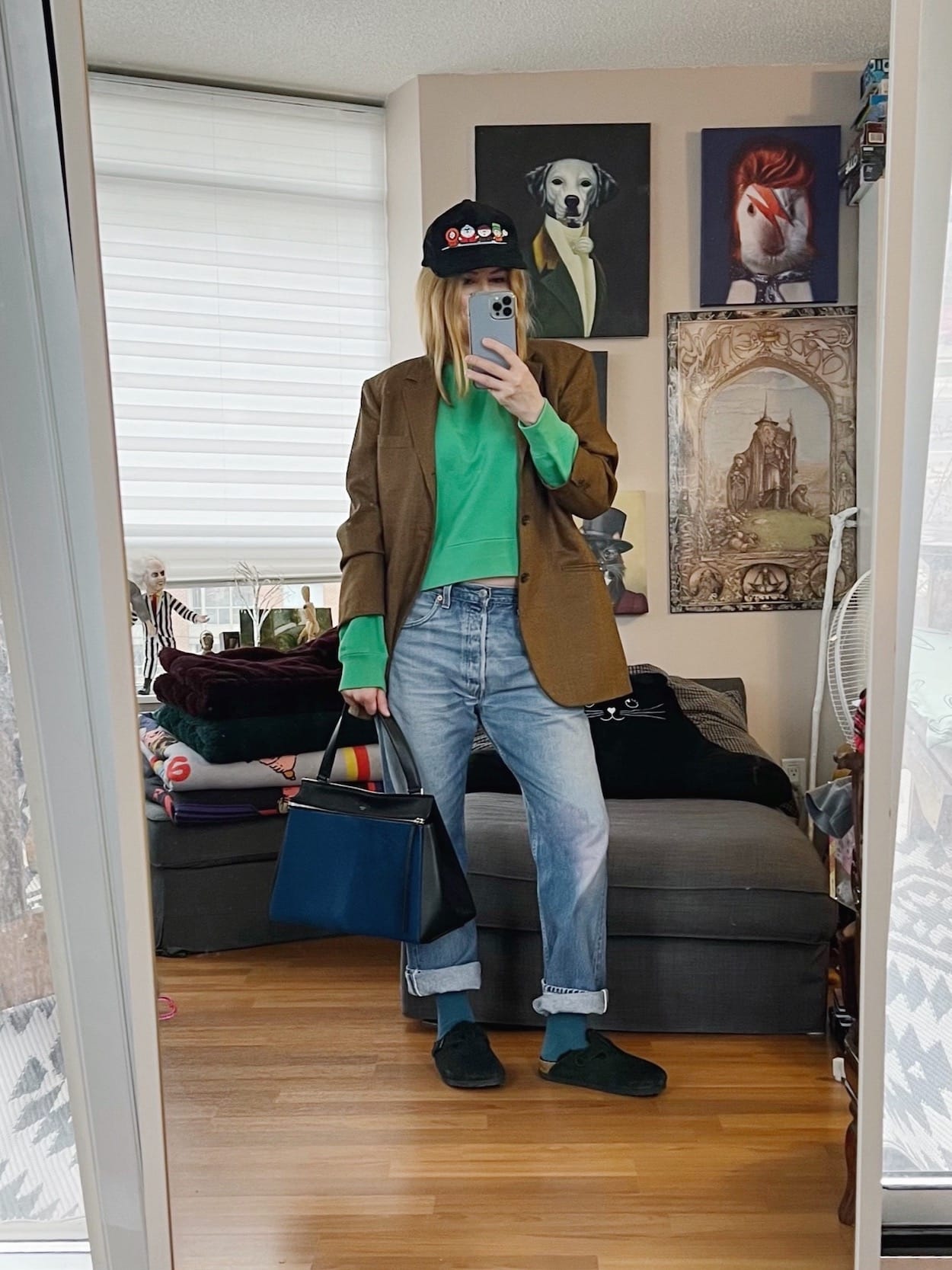 A blonde woman is wearing a green sweatshirt, vintage Levis', an oversized blazer, Birkenstock Bostons, a South Park cap, and a Celine bag.