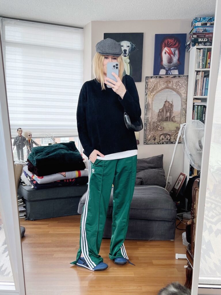 A blonde woman is wearing a black cashmere sweater with a white tee layered under, green Adidas track pants, Onituska Tigers, a driving cap, and a vintage Gucci bag.