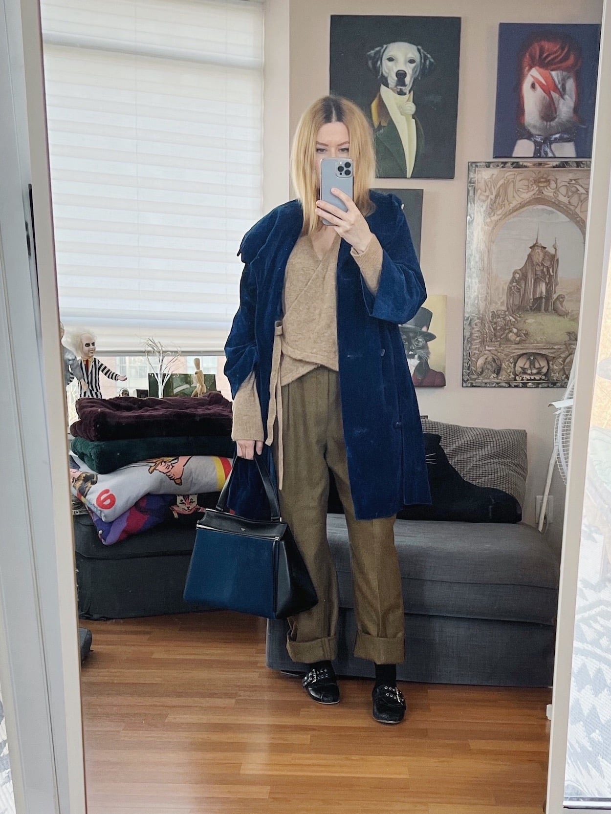 A blonde woman is wearing a wrap cardigan, vintage trousers, ballet flats, a vintage blue velvet jacket, and a Celine bag.