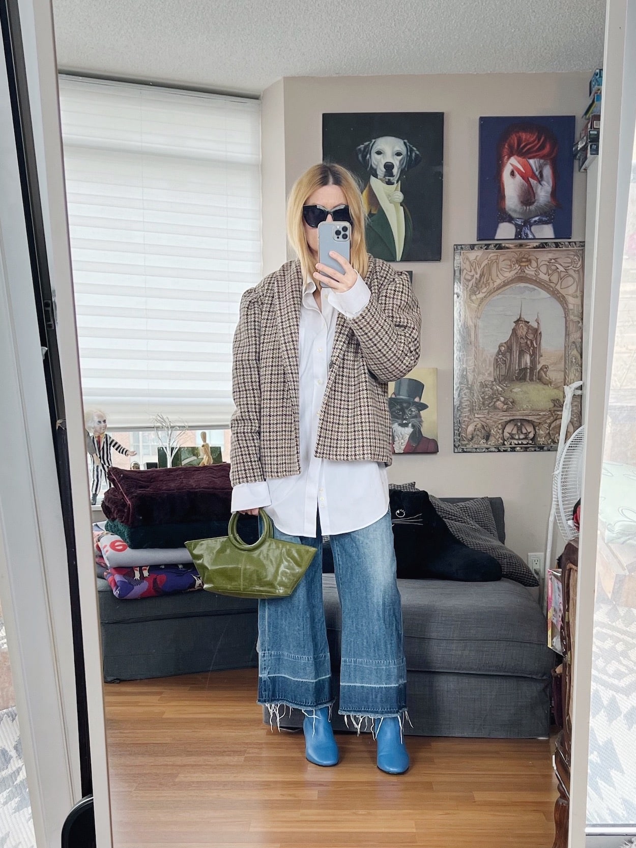 A blonde woman is wearing a white button up, frayed jeans, a houndstooth jacket, blue boots, and a vintage bag.