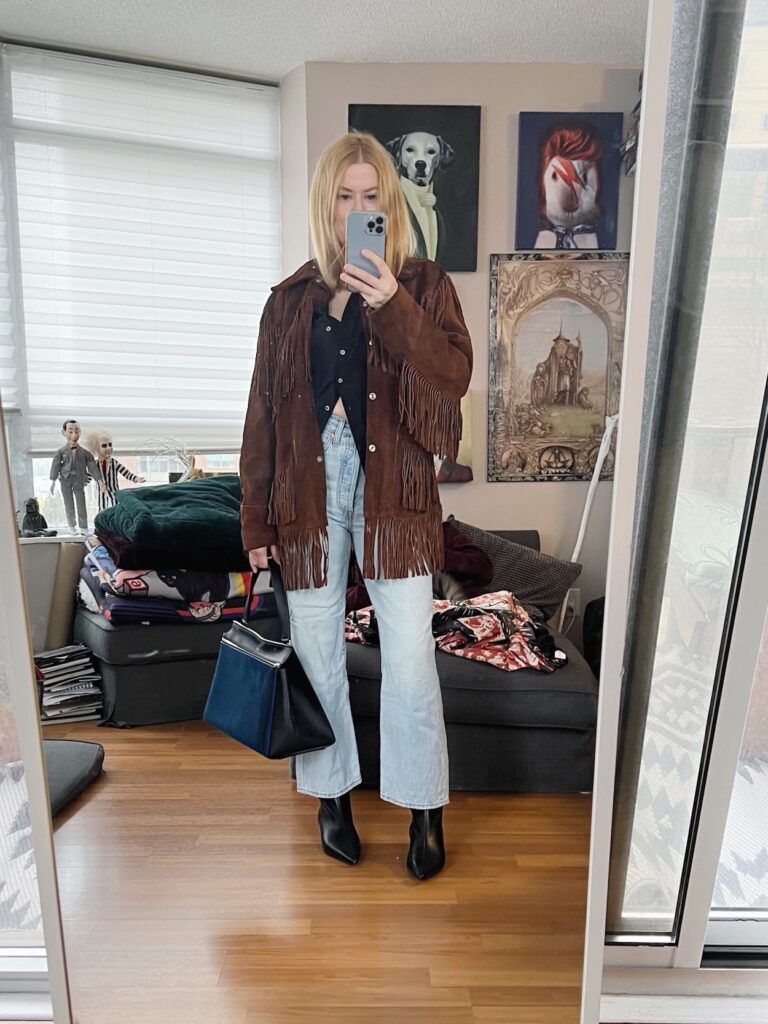 A blonde woman is wearing a black silk blouse, Levi's Rib cage, a vintage fringe jacket, and a Celine Edge bag.
