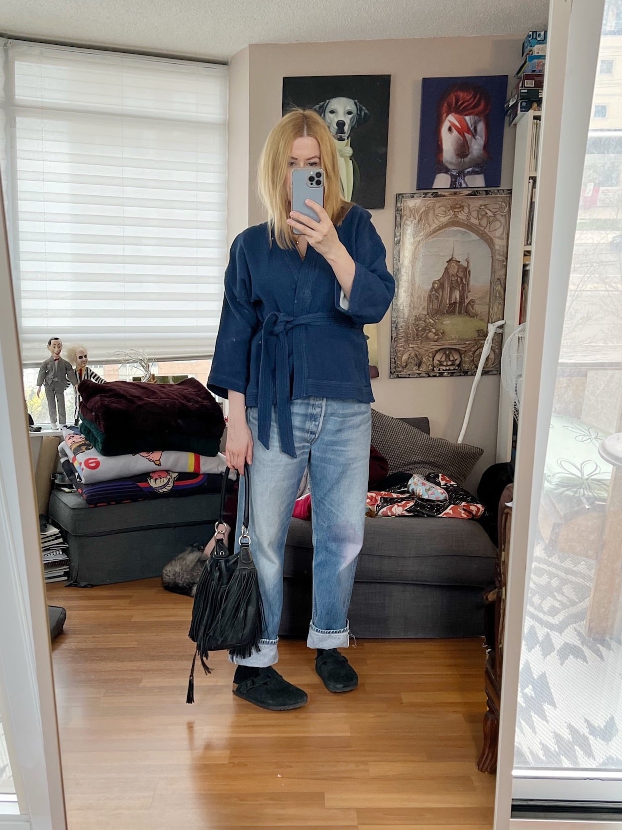 A blonde woman is wearing a blue wrap shirt, vintage Levi's, Birkenstock Boston clogs, and a fringe bucket bag.