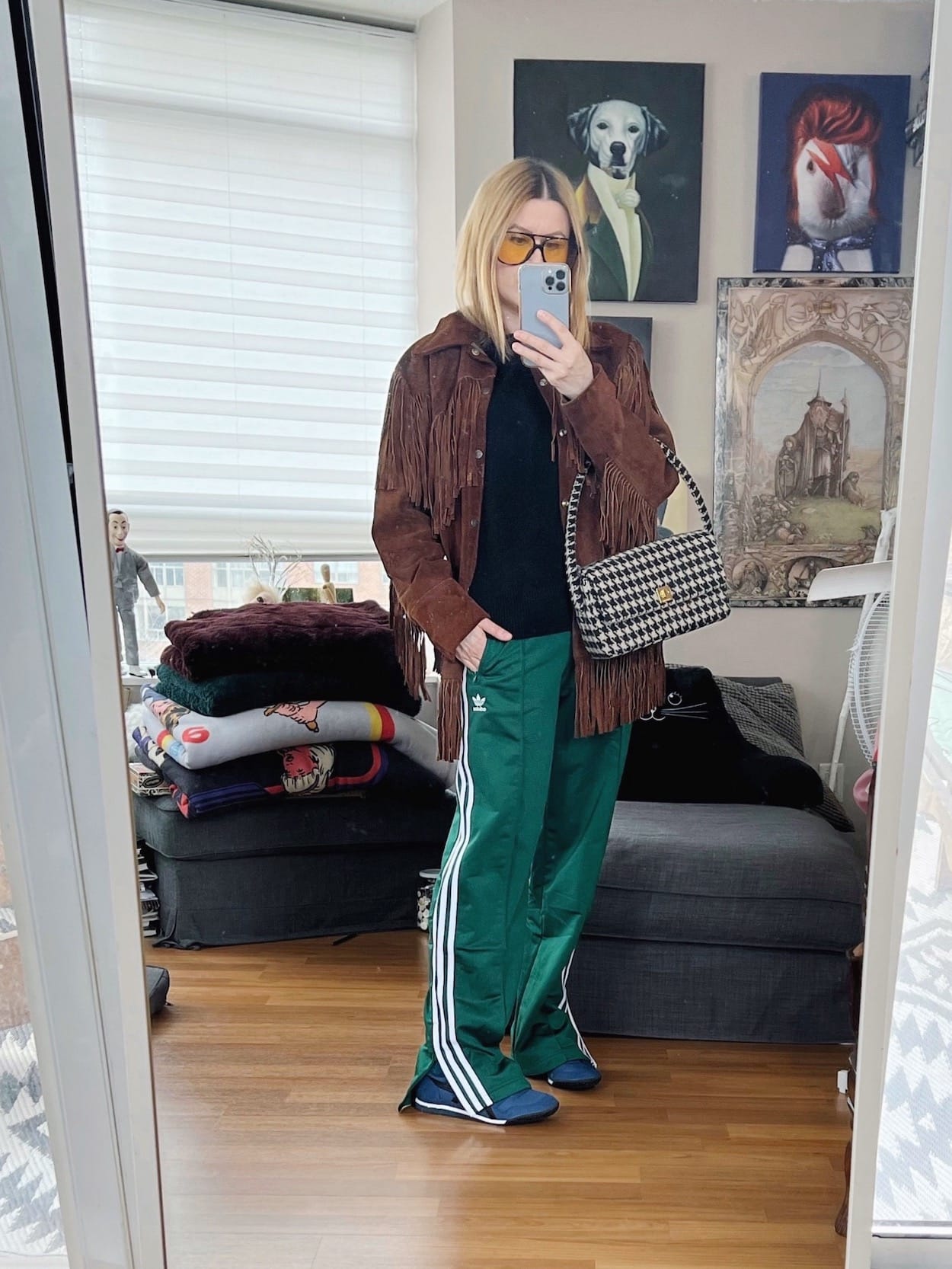 A blonde woman is wearing a vintage fringe jacket, Adidas Firebird track pants, a black sweater, Onitsuka sneakers, retro sunglasses, and an Anine Bing bag.