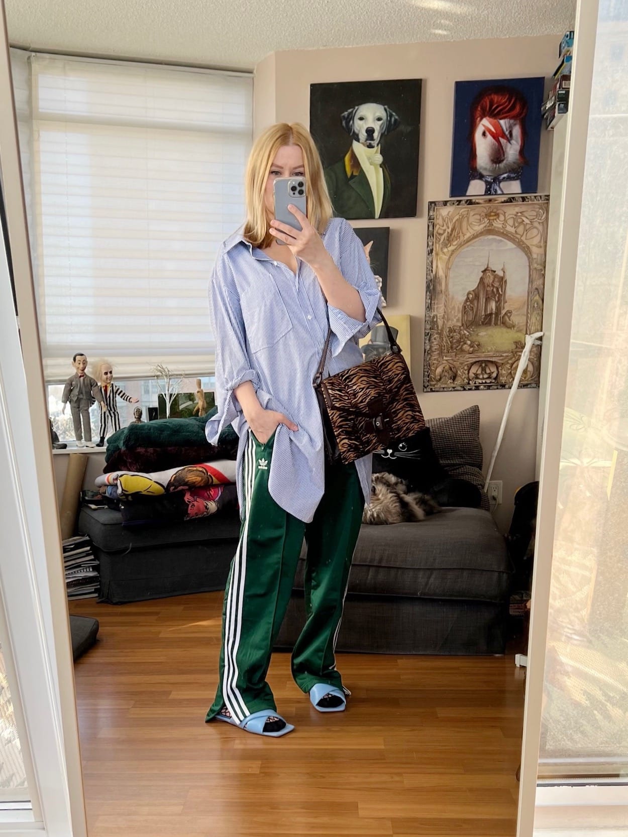 A blonde woman is wearing an oversized striped button up, green Adidas Firebird pants, blue sandals, and a vintage animal print bag.