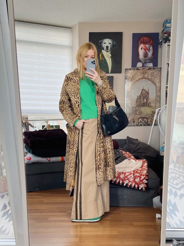 A blonde woman is wearing a green sweatshirt, wide leg trousers, a vintage animal print coat, green New Balance sneakers, and a vintage Gucci bag.