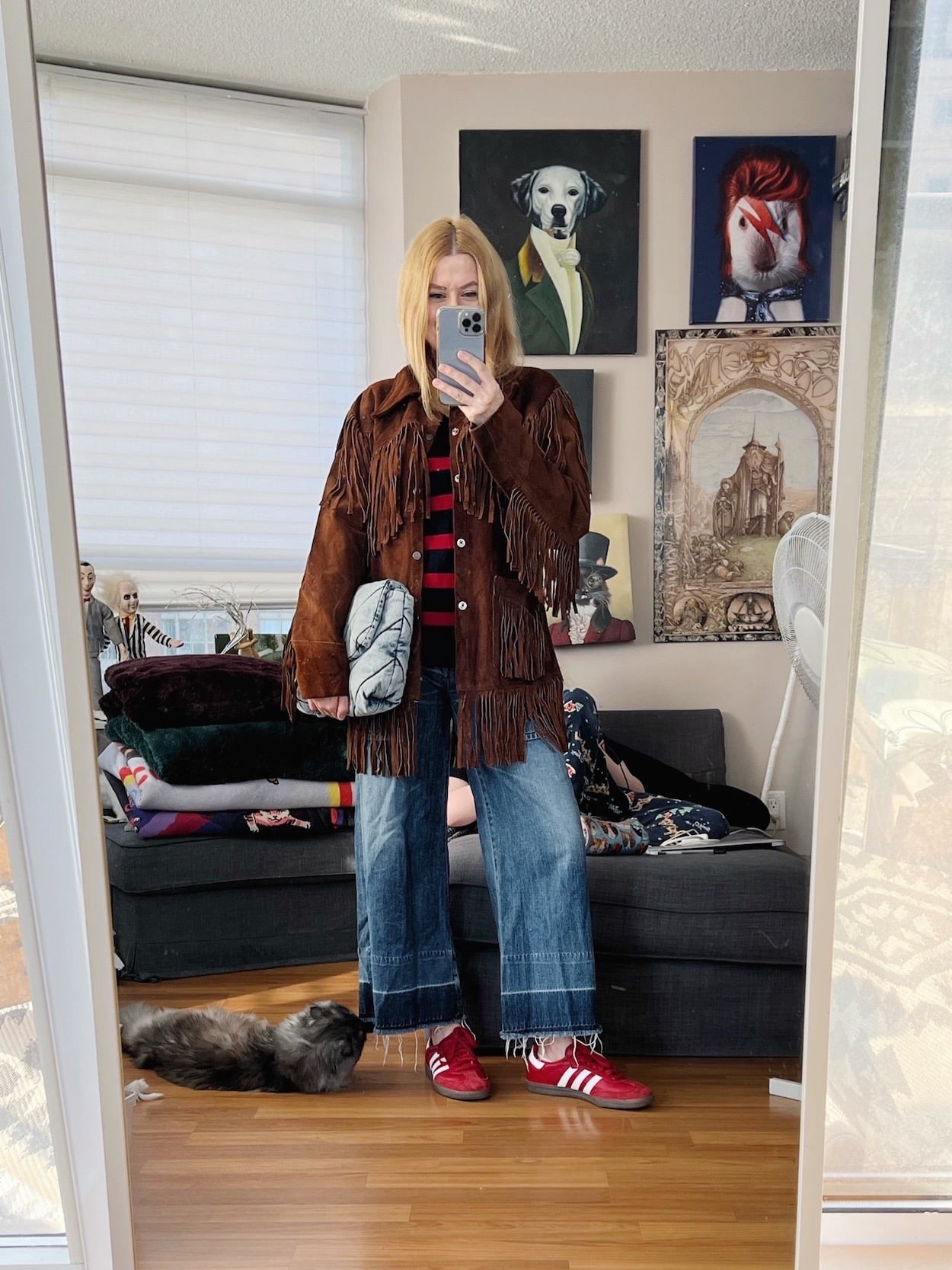 A blonde woman is wearing a striped sweater, cropped jeans, red Adidas Sambas, a vintage fringe jacket, and a denim clutch.