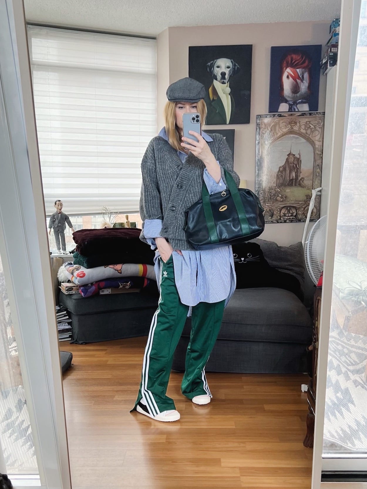 A blonde woman is wearing Adidas Firebird track pants, an oversized button up, an asymmetrical cardigan, a driving cap, and a vintage Longchamp bag.