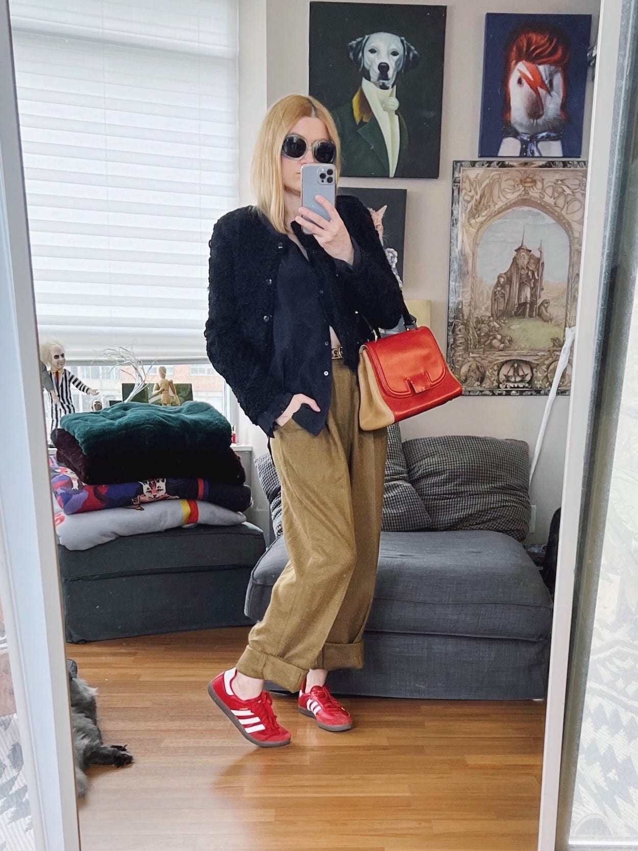 A blonde woman is wearing a black silk blouse, black cropped jacket, vintage trousers, red Sambas, vintage sunglasses, and a Fendi handbag.