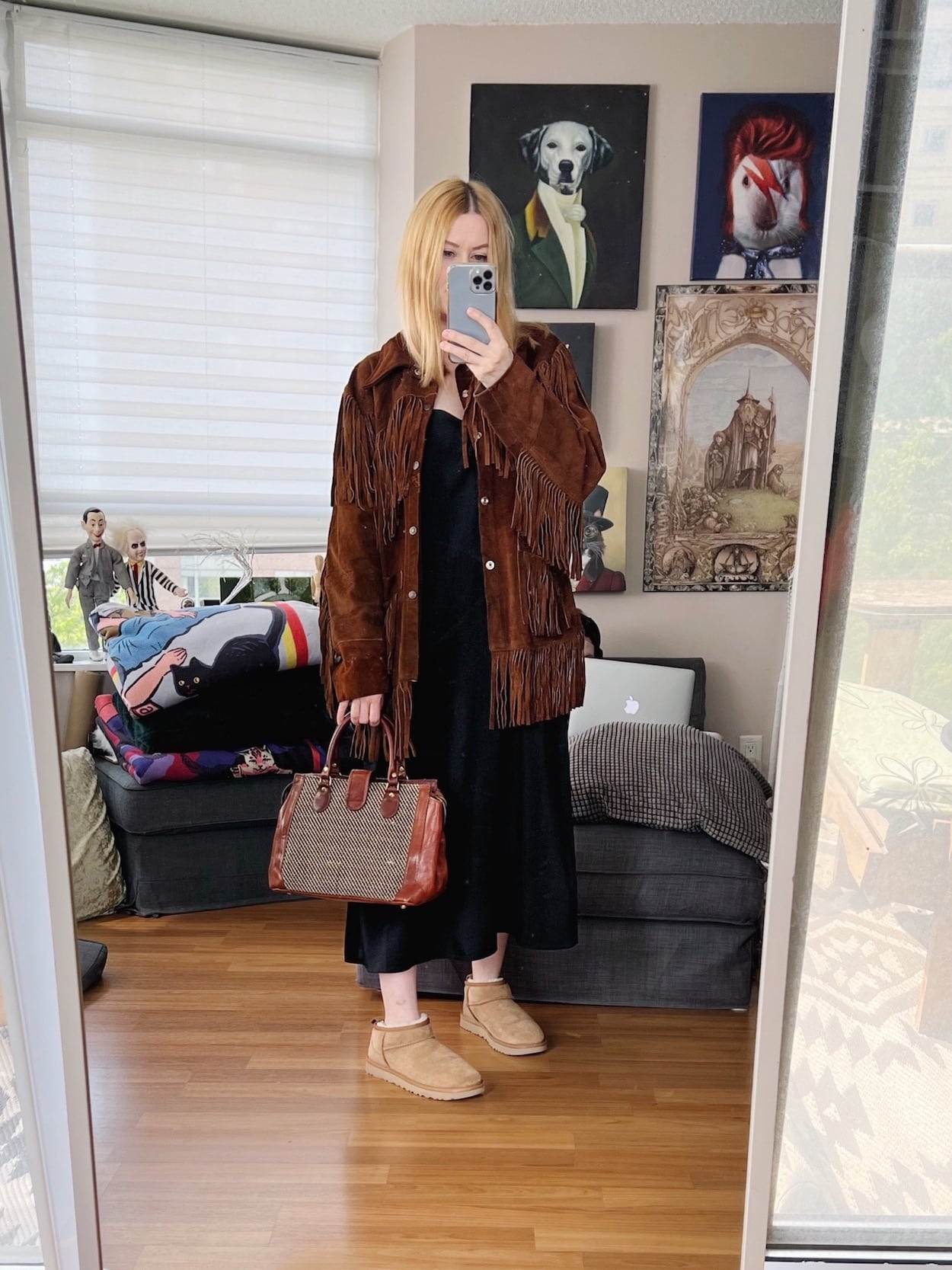 A blonde woman is wearing a slip dress, vintage fringe jacket, ugg booties, and carrying a vintage handbag.