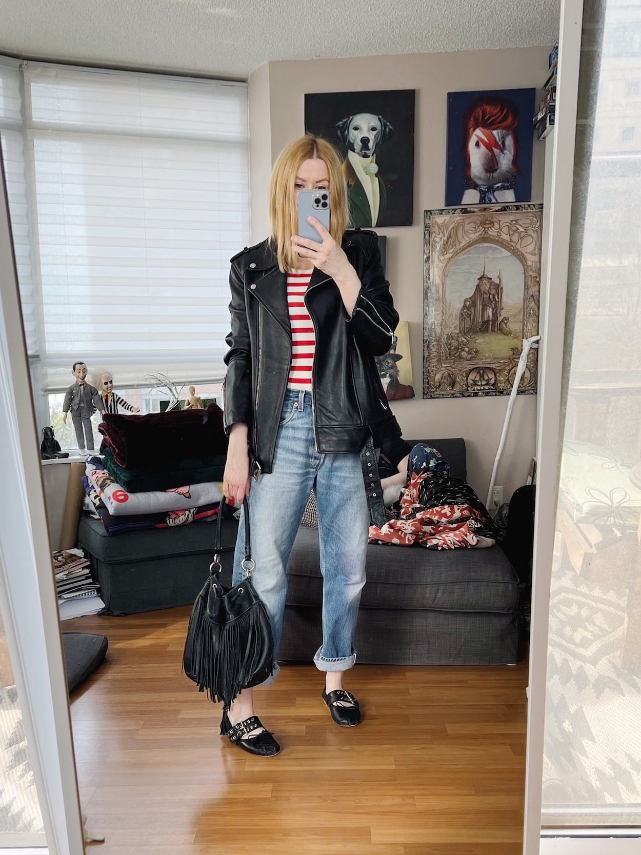 A blonde woman is wearing a striped tank top, vintage Levi's an oversized leather moto, Miu Miu ballet flats, and a vintage fringe bucket bag.