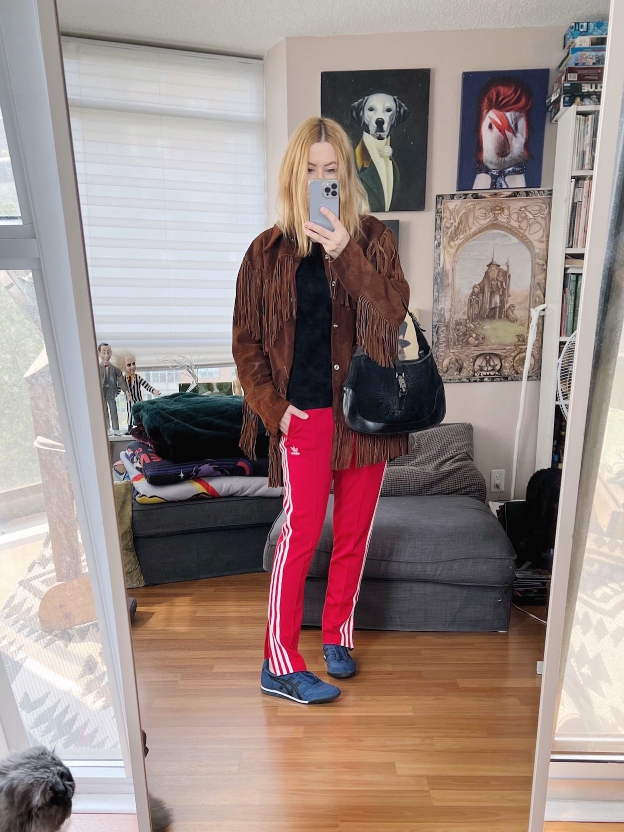 A blonde woman is wearing a black cashmere sweater with red Adidas track pants, a vintage fringe jacket, blue Onitsuka Tigers, and a vintage Gucci bag,