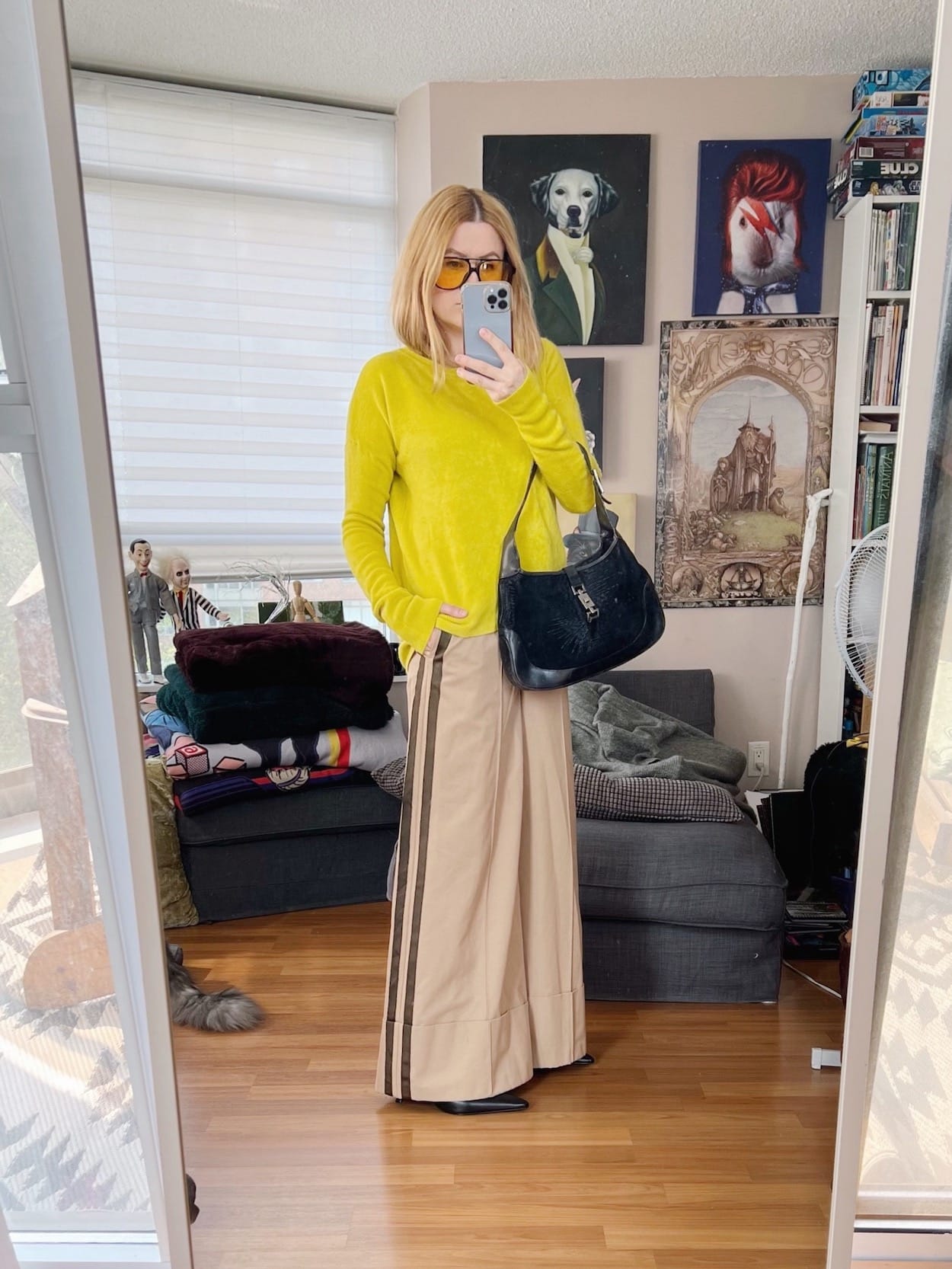 A blonde woman is wearing a yellow sweater, wide leg trousers, pointy toed boots, retro sunglasses, and a vintage Gucci bag.