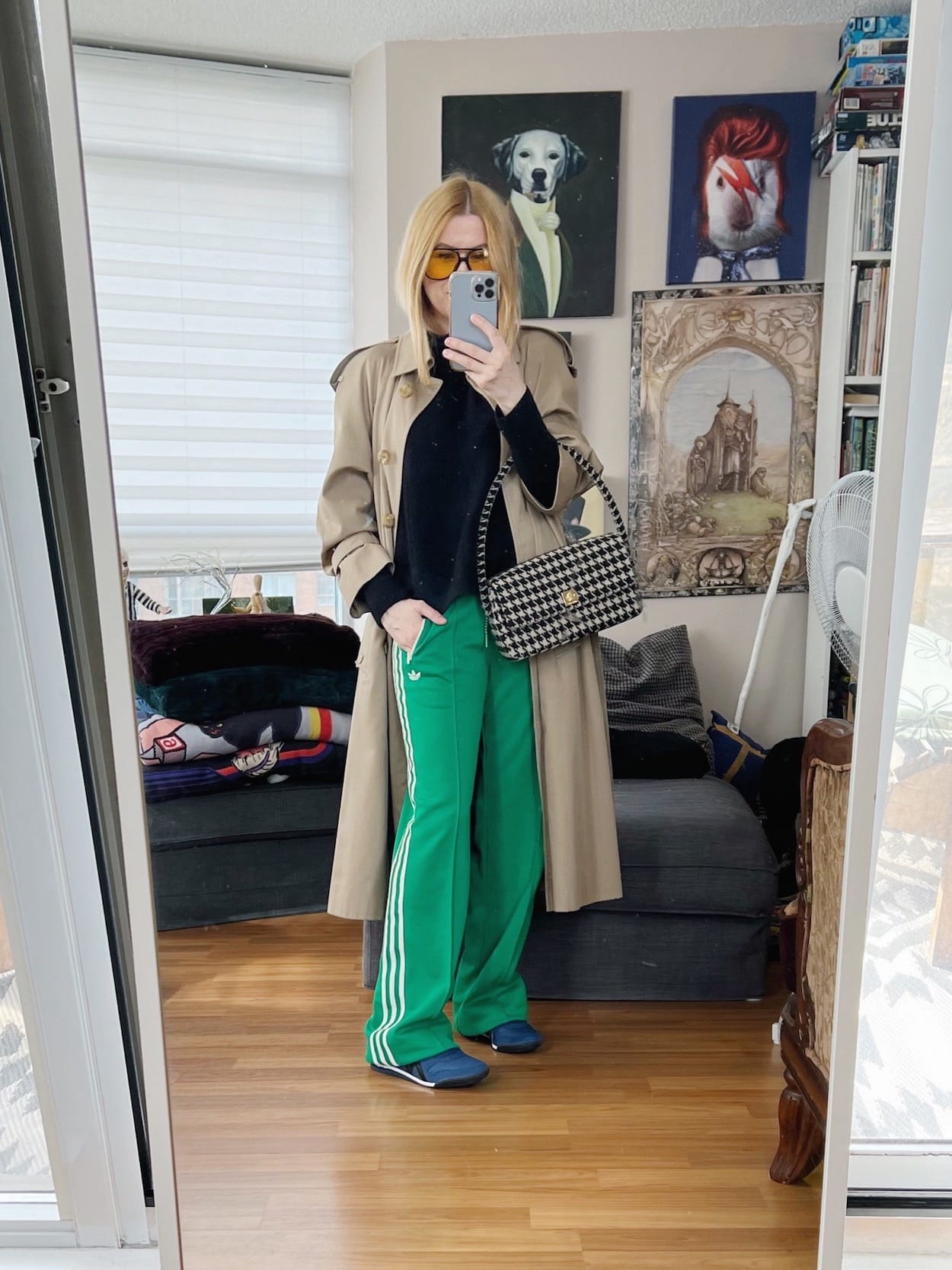 A blonde woman is wearing green Adidas track pants, a black sweater, a vintage trench coat, Onitsuka Tiger sneakers, retro sunglasses, and an Anine Bing houndstooth handbag.