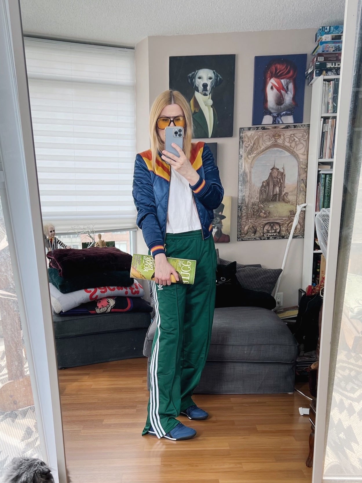 A blonde woman is wearing a white t-shirt, a classic rock couture bomber jacket, Adidas Firebird track pants, Onitsuka Tiger sneakers, retro sunglasses, and a 1970s magazine clutch.
