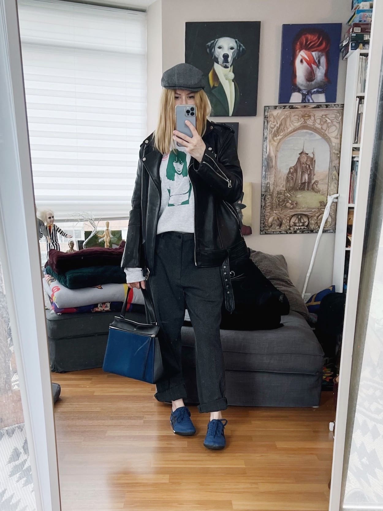 A blonde woman is wearing a Bjork sweatshirt, grey trousers, and oversized leather moto, Onitsuka Tiger sneakers, a hat, and a Celine Edge bag.