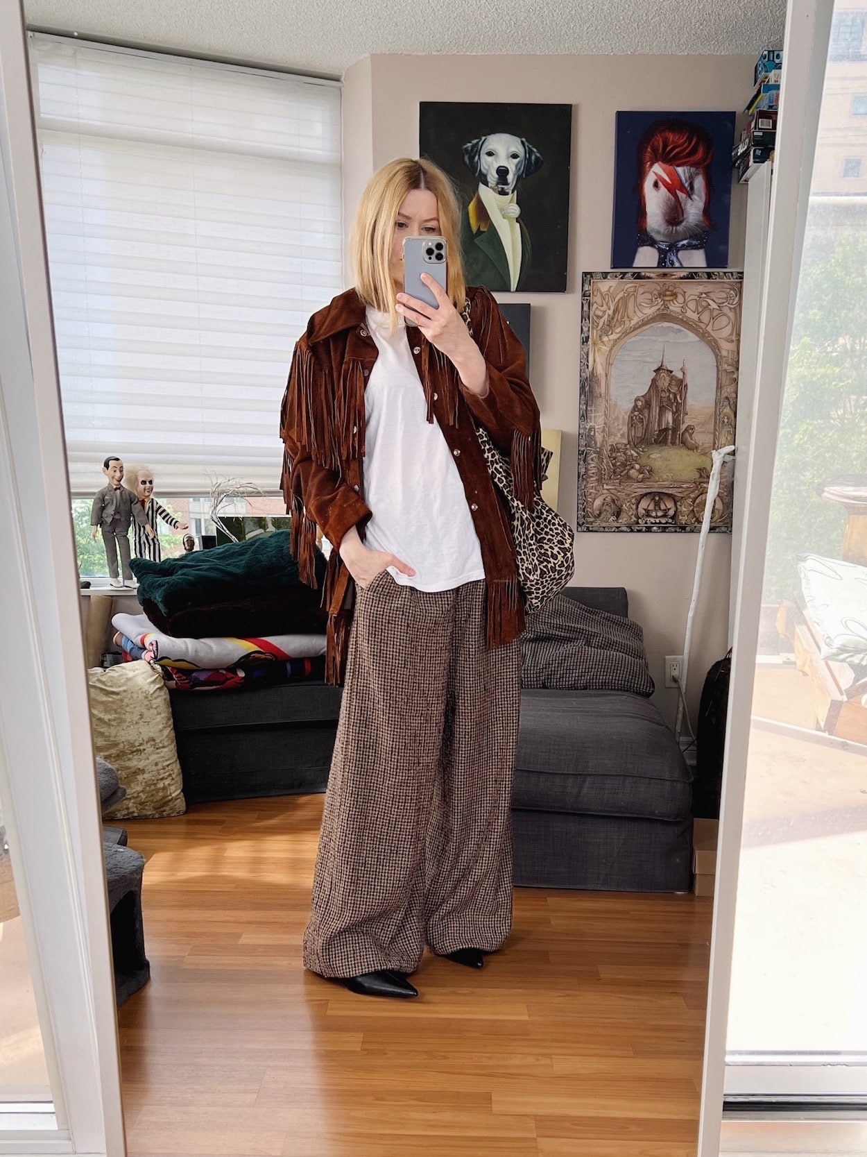 A blonde woman is wearing wide leg trousers, a white t-shirt, a vintage fringe jacket, kitten heel boots, and an animal print tote.