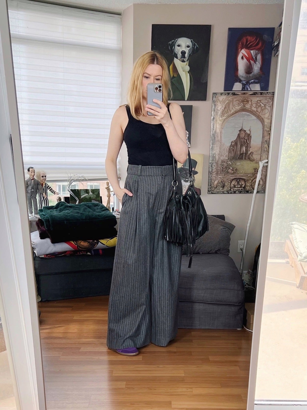 A blonde woman is wearing a black tank, wide leg grey trousers, purple sneakers, and a vintage fringe bag.