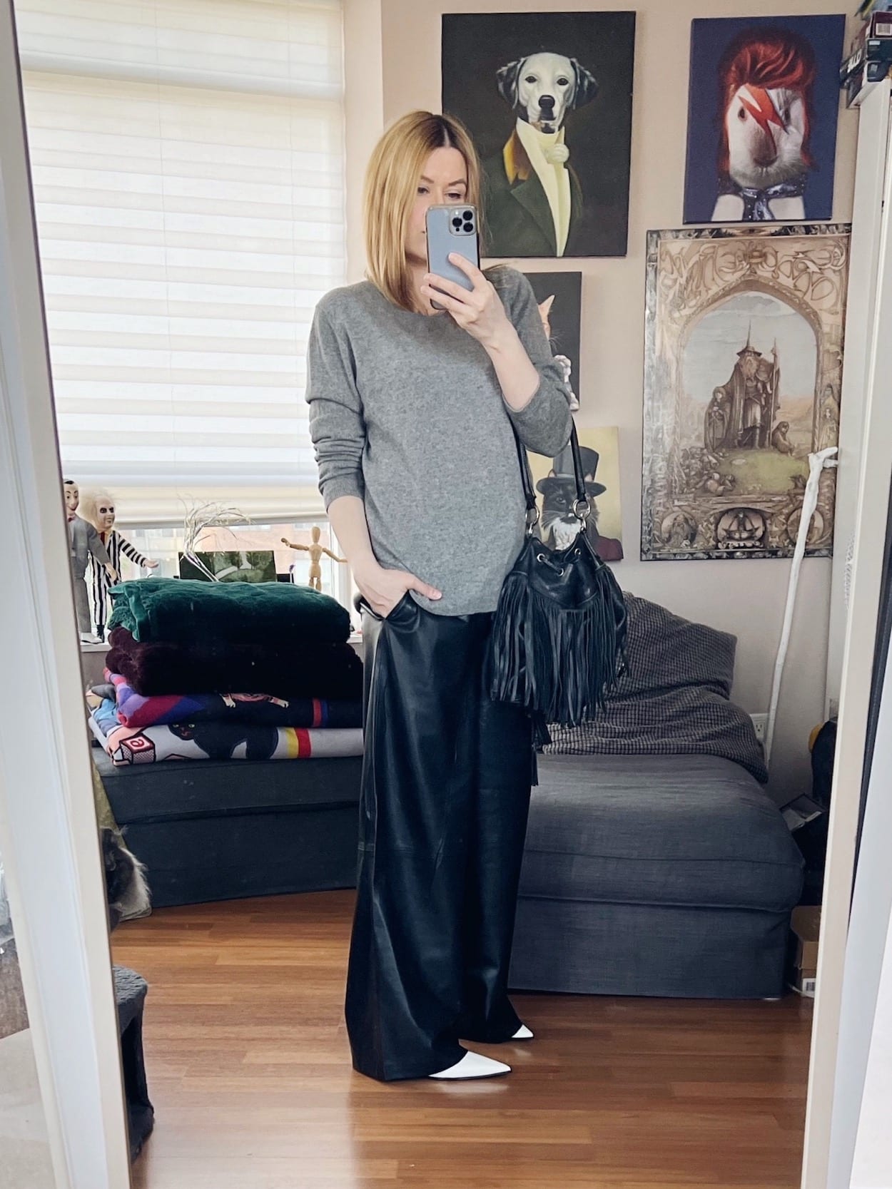 A blonde woman is wearing a grey cashmere sweater, wide leg leather trousers, white boots, and a vintage fringe bucket bag.