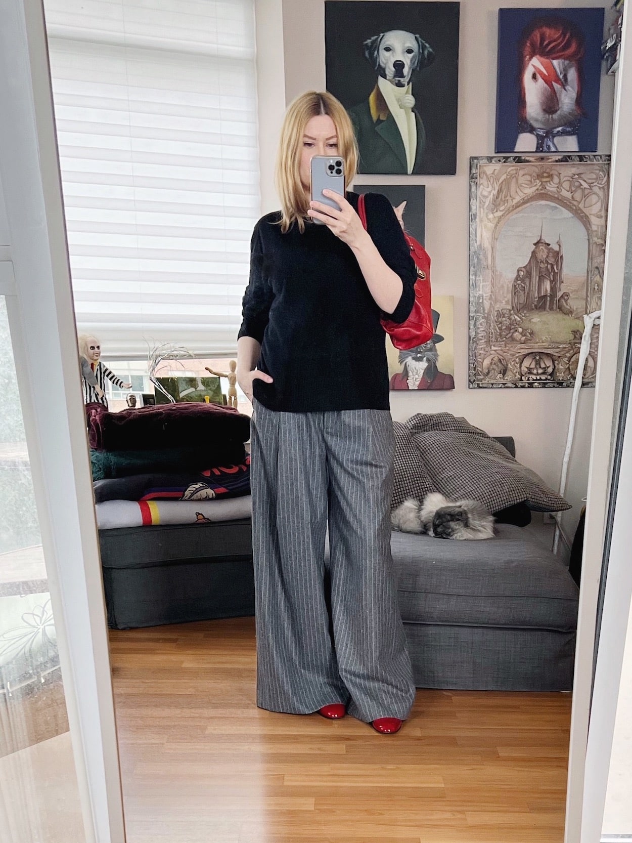 A blonde woman is wearing grey wide leg trousers, a black cashmere sweater, red Mary Janes, and a red Prada handbag.