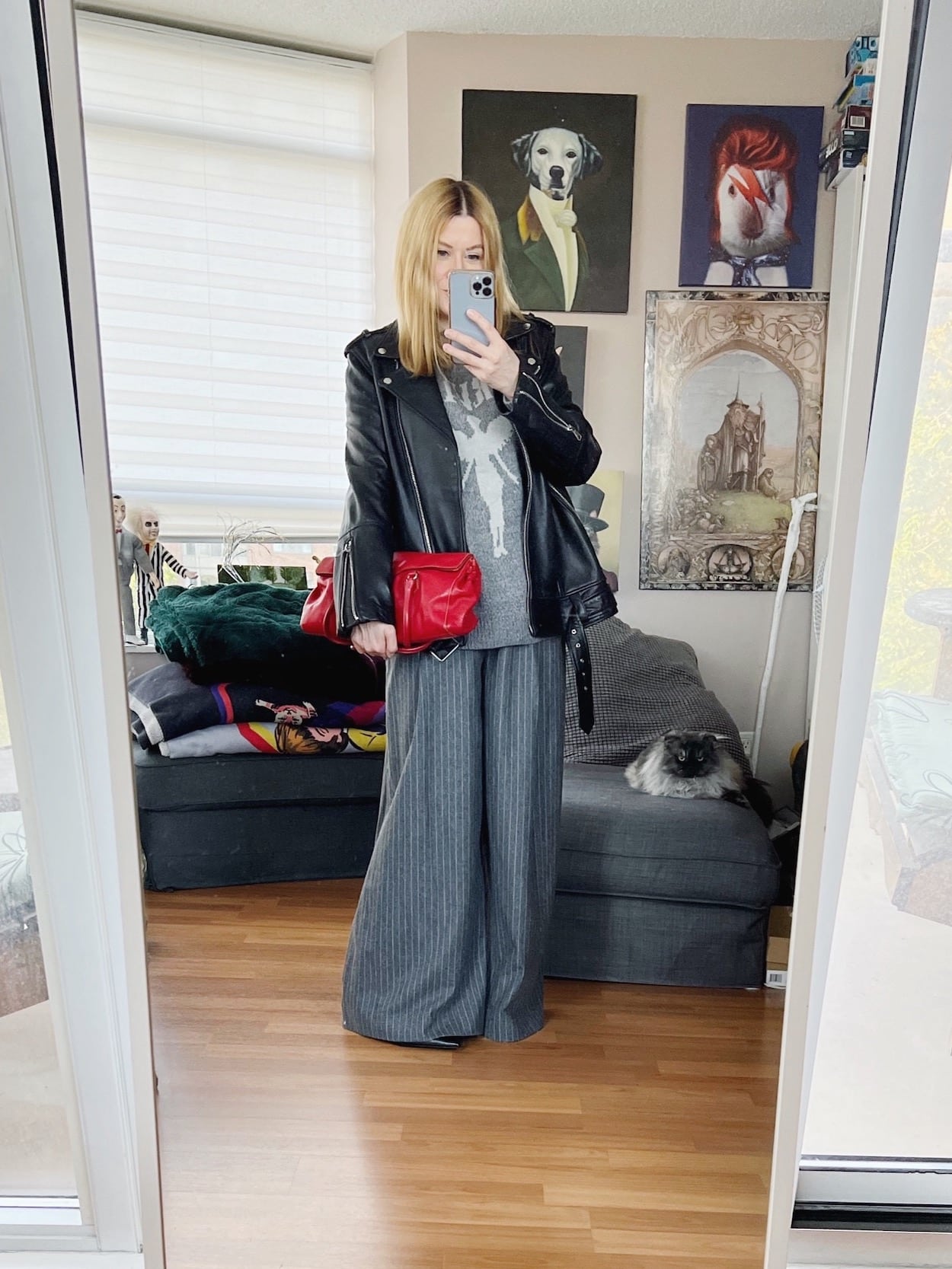 W blonde woman is wearing wide leg trousers, a Nirvana sweater, an oversized moto jacket, black boots, and a red handbag.