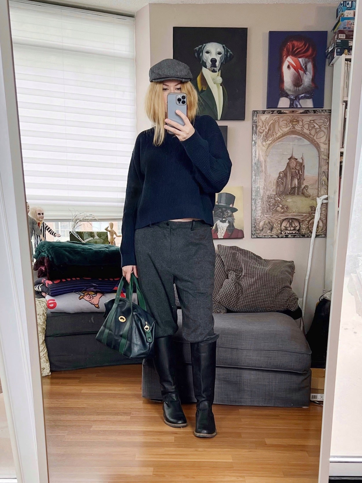 A blonde woman is wearing a navy blue sweater, men's trousers, riding boots, a flat cap, and a vintage Longchamp handbag.