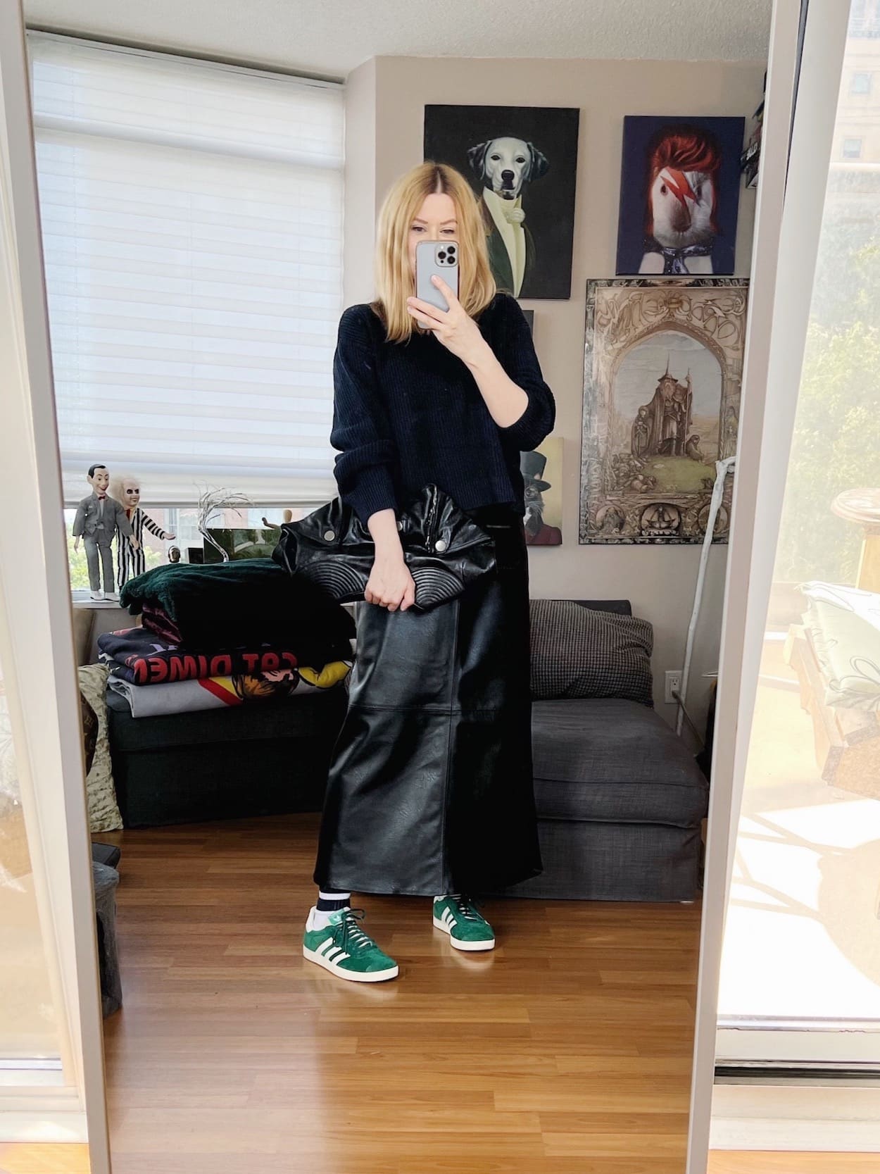 A blonde woman is wearing a navy blue sweater, black faux leather maxi skirt, green Adidas, and an Alexander McQueen clutch.