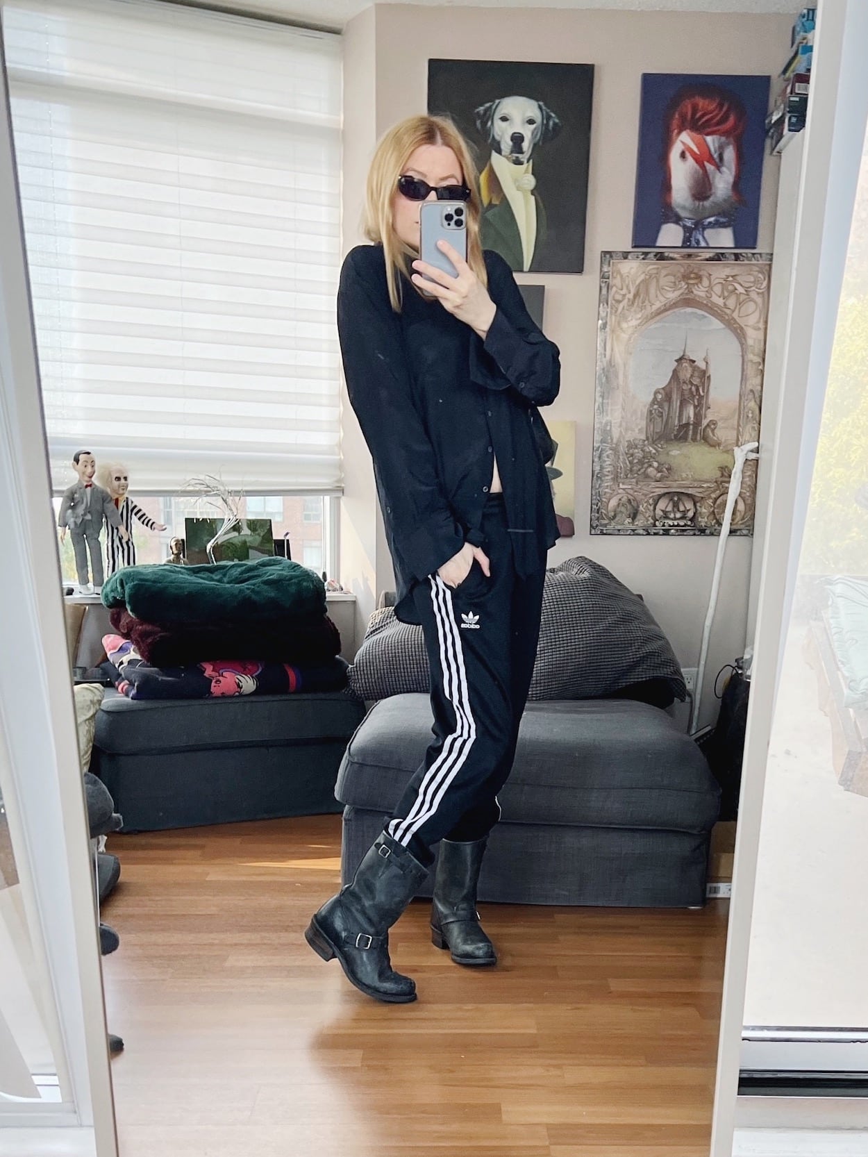 A blonde woman is wearing a black silk blouse, black Adidas track pants, Frye boots, a vintage bag, and cat eye sunglasses.