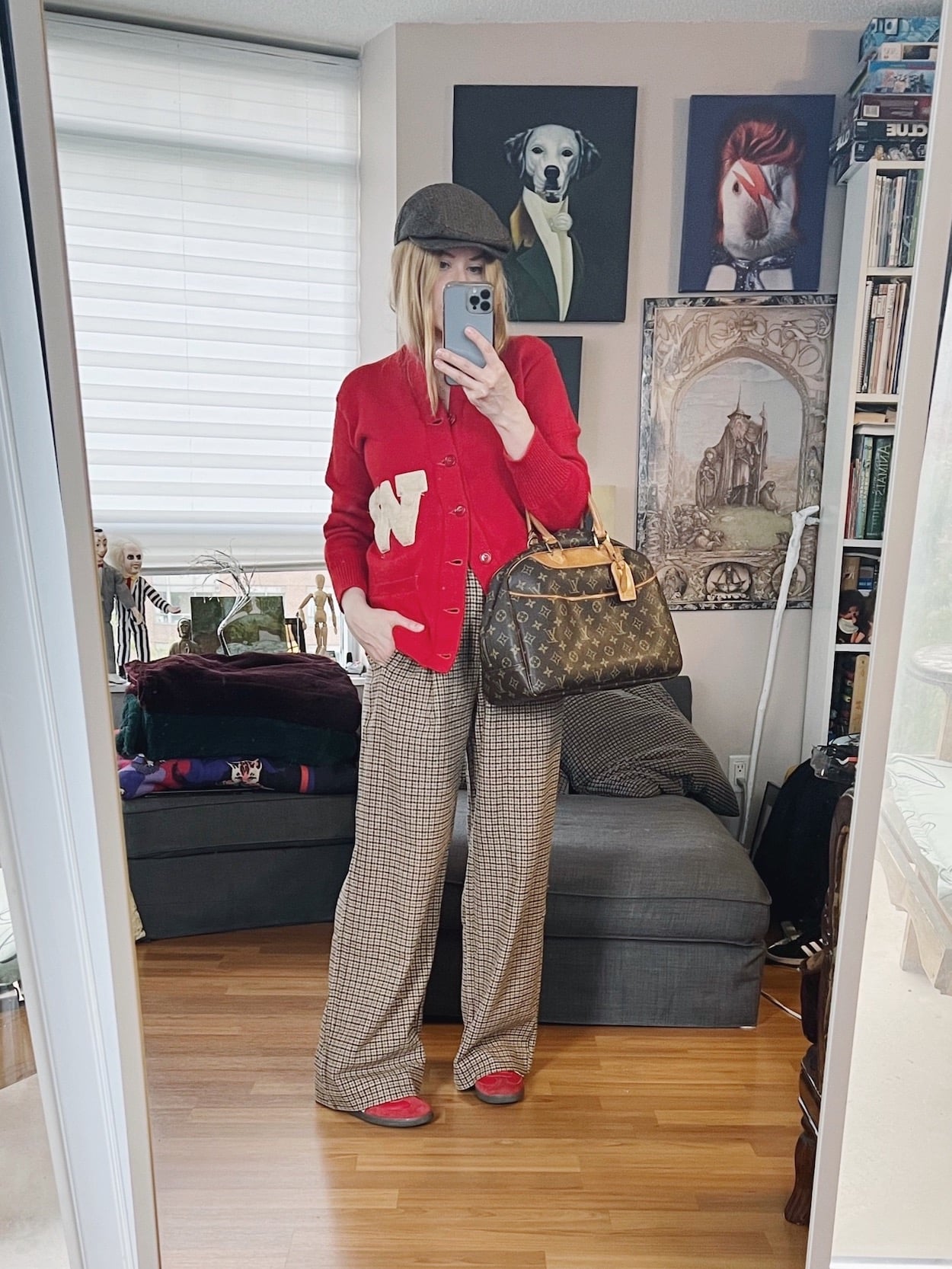 A blonde woman is wearing a vintage letterman cardigan, houndstooth trousers, red Sambas, a flat cap, and is carrying a vintage Louis Vuitton bag.