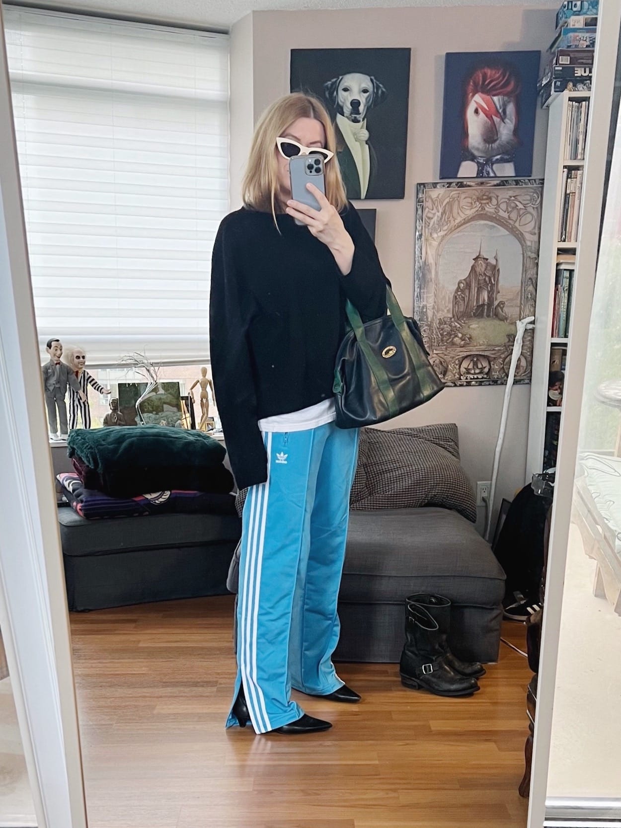 A blonde woman is wearing a black cashmere sweater layered over a white tee, blue Adidas track pants, kitten heel boots, white Celine sunglasses, and is carrying a vintage Longchamp bag.