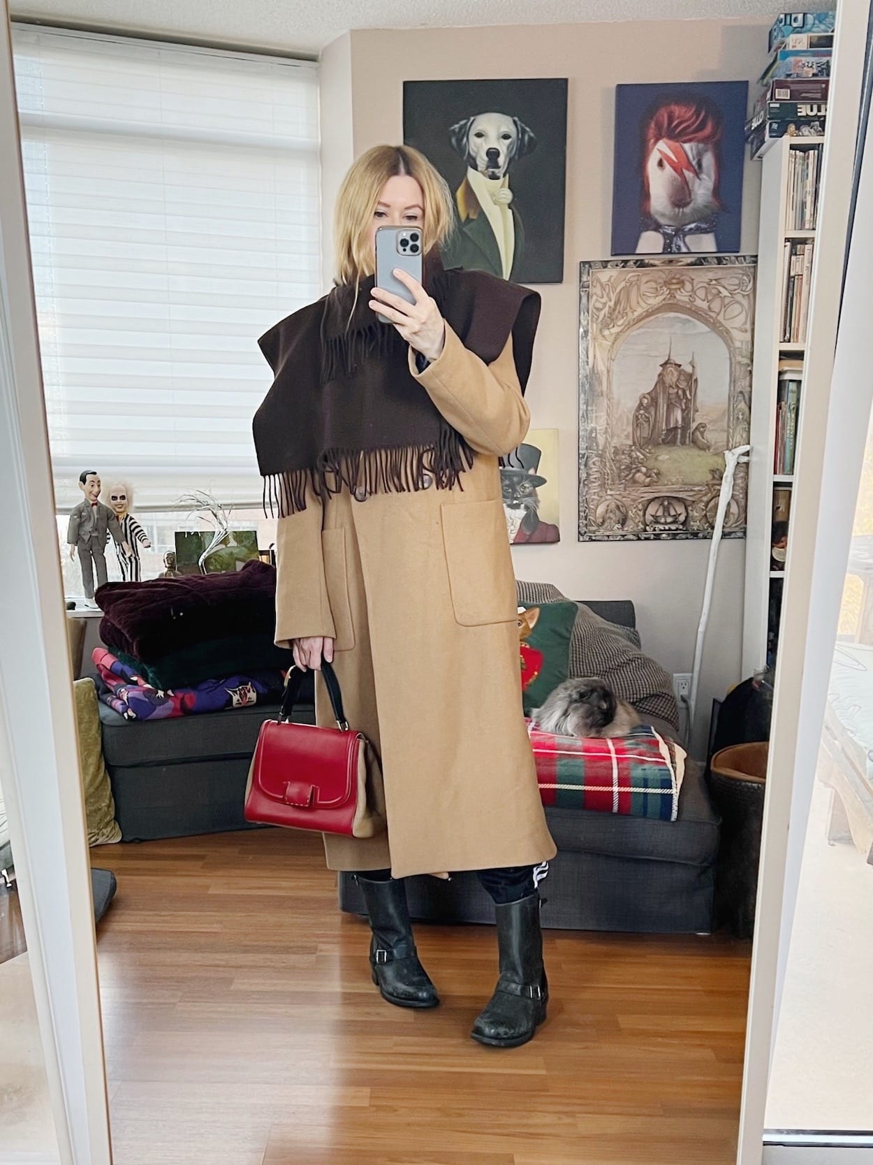 A blonde woman is wearing a adidas track pants, a sweater, under a long camel coat, moto boots, a fringed short cape, and is carrying a vintgae Fendi bag.