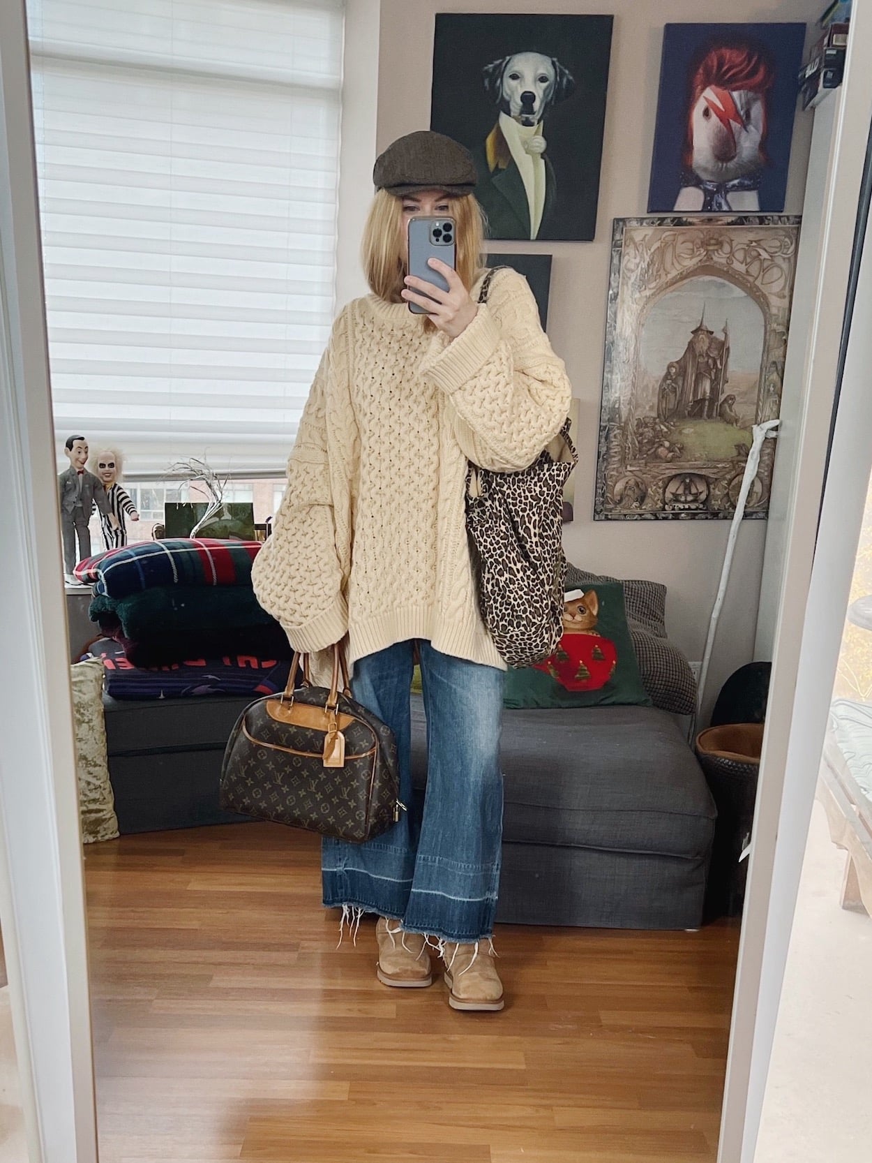 A blonde woman is wearing an oversized cable knit sweater, wide leg jeans. Mini Uggs, a flat cap, and is carrying an animal print tote, and vintage Louis Vuitton handbag.