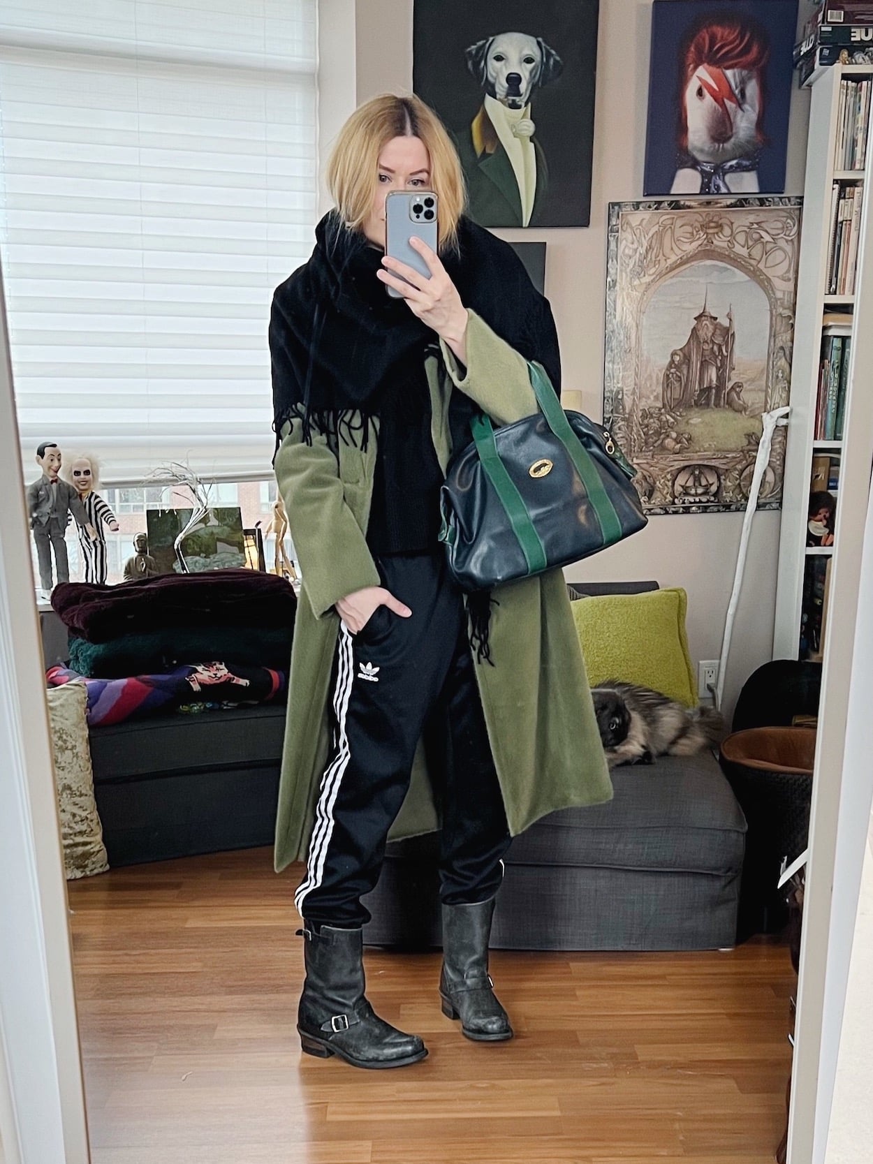 A blonde woman is wearing a black sweater, black Adidas track pants, moto boots, a green coat with a black shawl scarf and is carrying a vintage bag.
