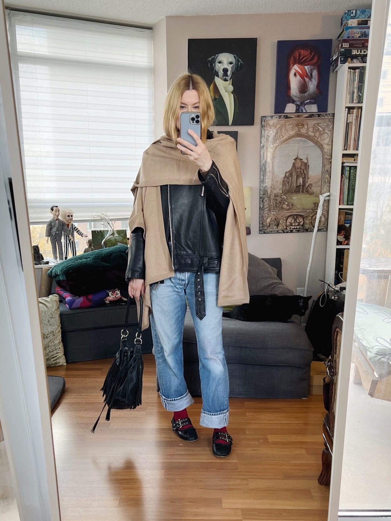 A blonde woman is wearing vintage jeans, a turtleneck, oversized moto jacket, a cape, red socks, ballet flats, and a vintage fringe bucket bag.