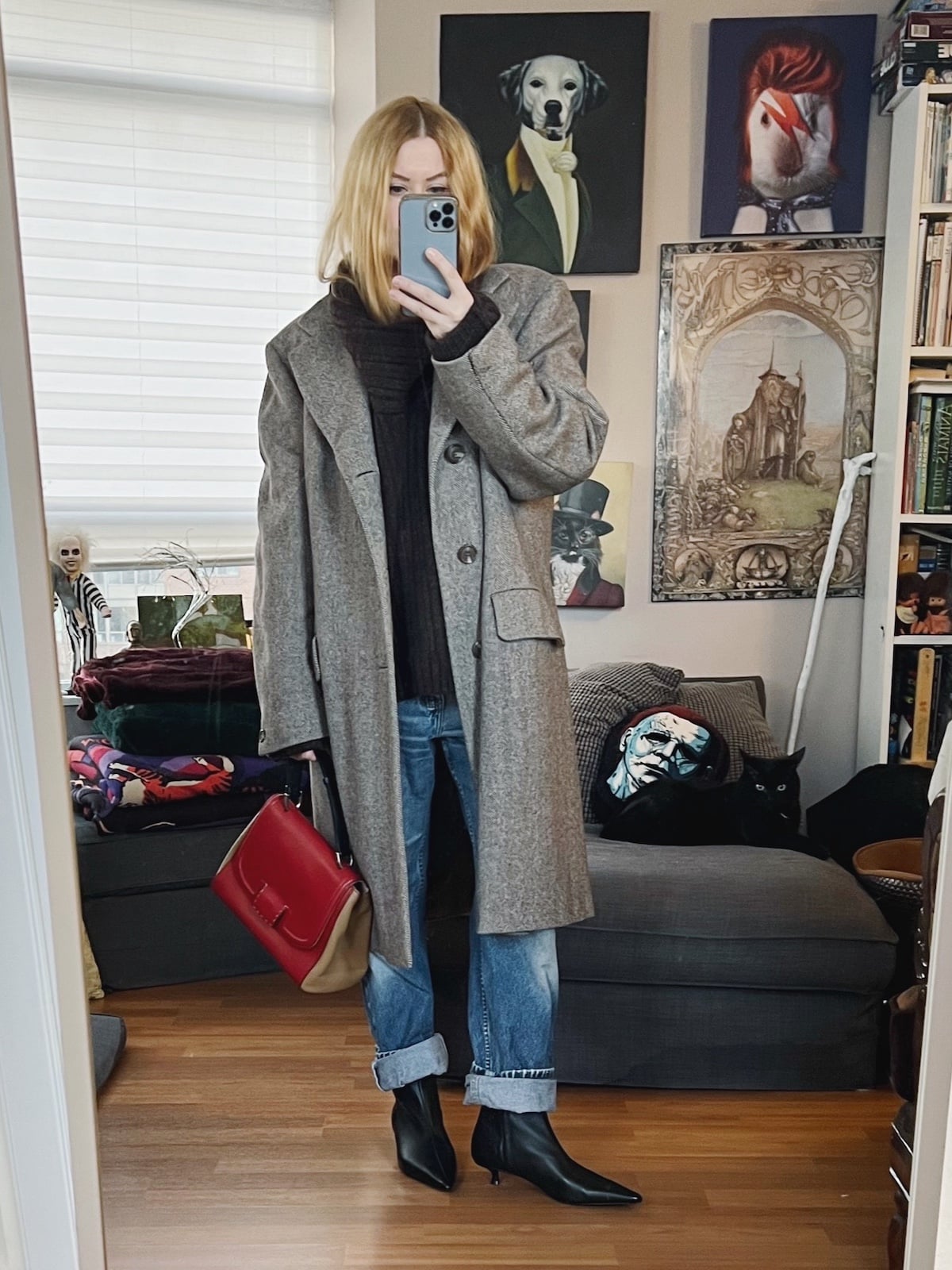 A blonde woman is wearing a brown sweater, vintage jeans, black boots, a men's vintage coat, and an old Fendi handbag.