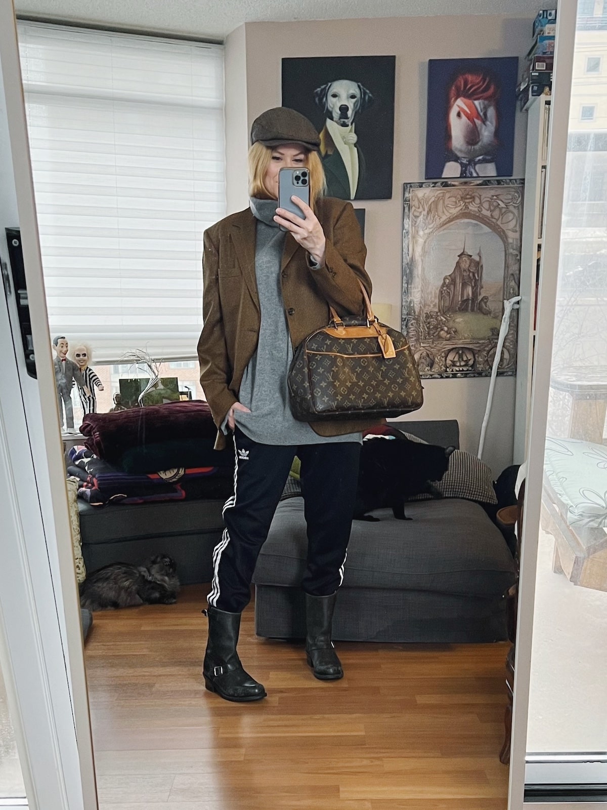 A blonde woman is wearing an oversized grey sweater, Adidas track pants, moto boots, a vintgae blazer, a flat cap, and a vintage Louis Vuitton bag. 