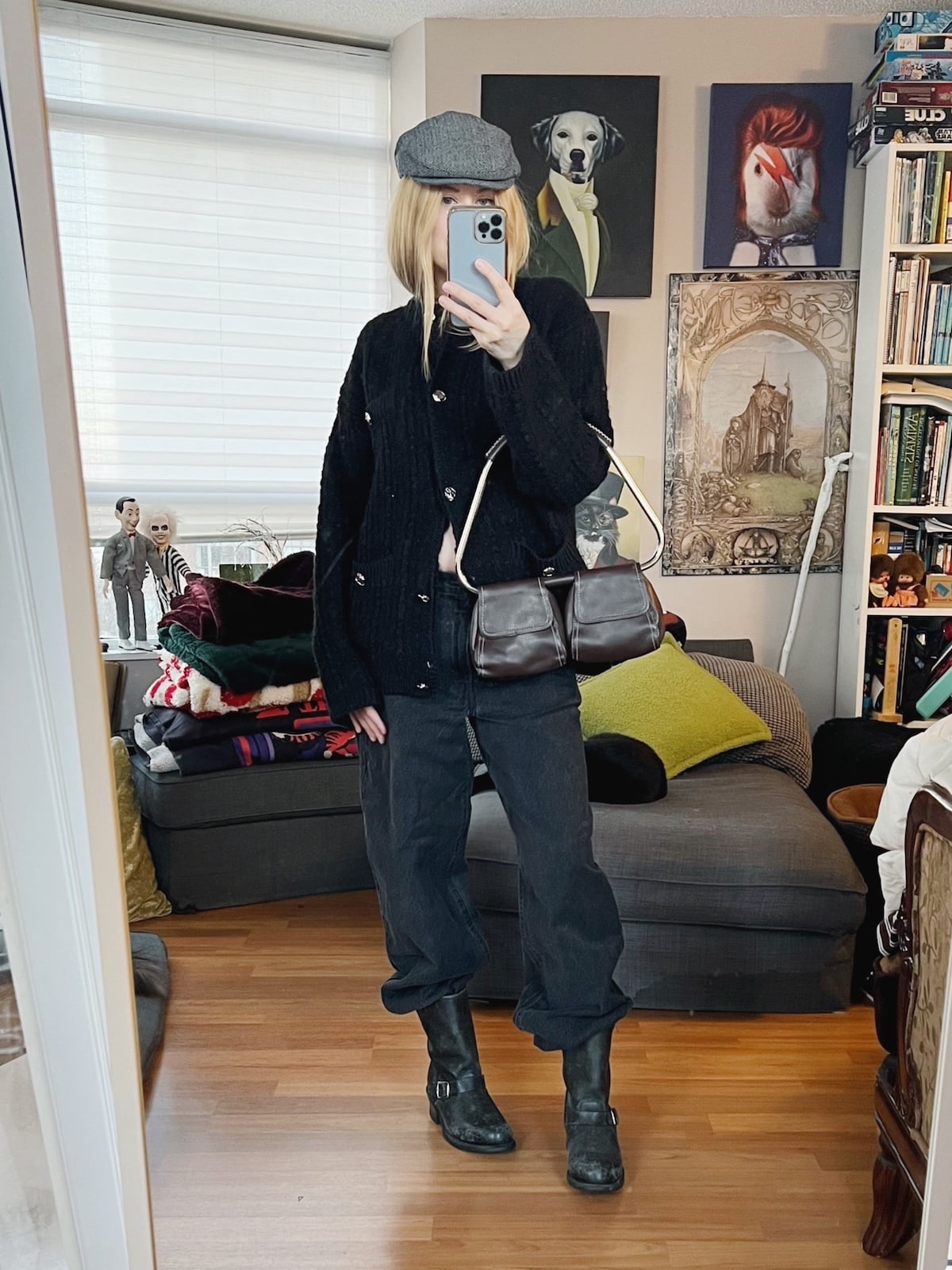 A blonde woman is wearing a black cardigan, black jeans, Frye moto boots, a flat cap and is carrying a vintage bag.
