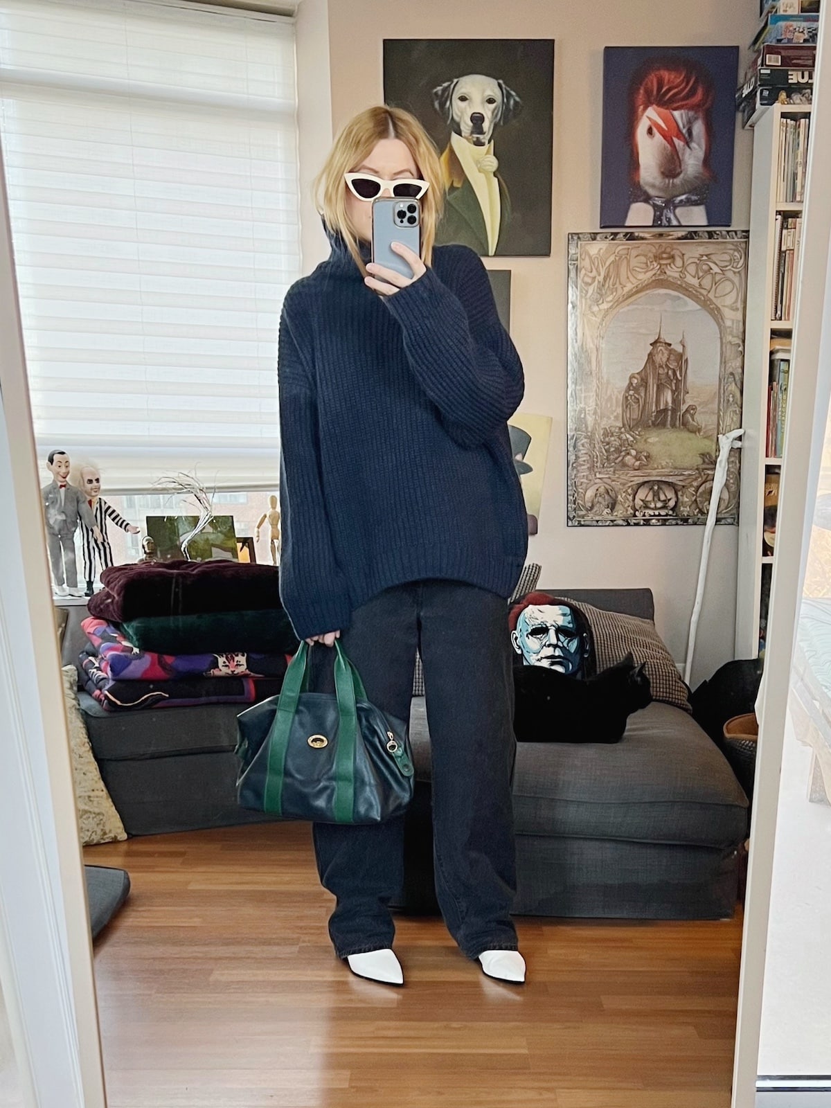A blonde woman is wearing a navy blue sweater, black jeans, white boots, white sunglasses, and a vintage bag.