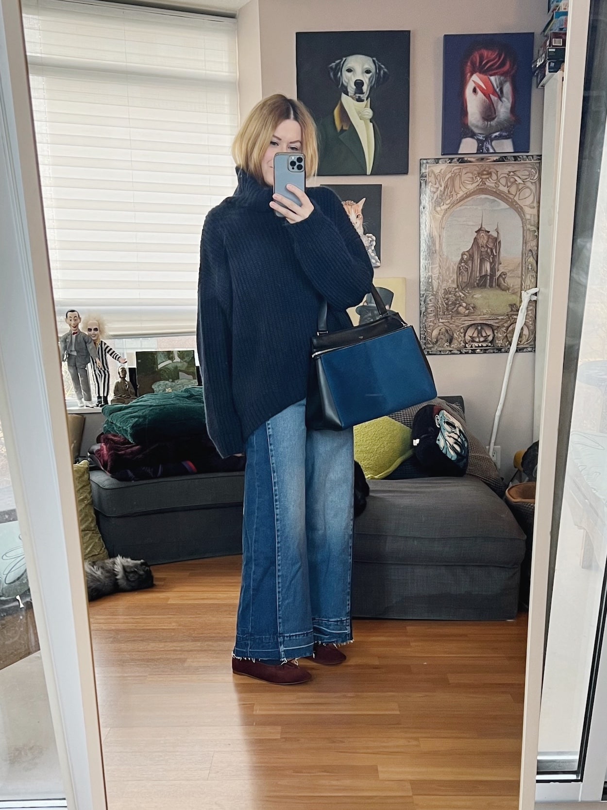 A blonde woman is wearing a navy blue oversized knit, wide leg jeans, brown ballet flats, and an old Celine bag.