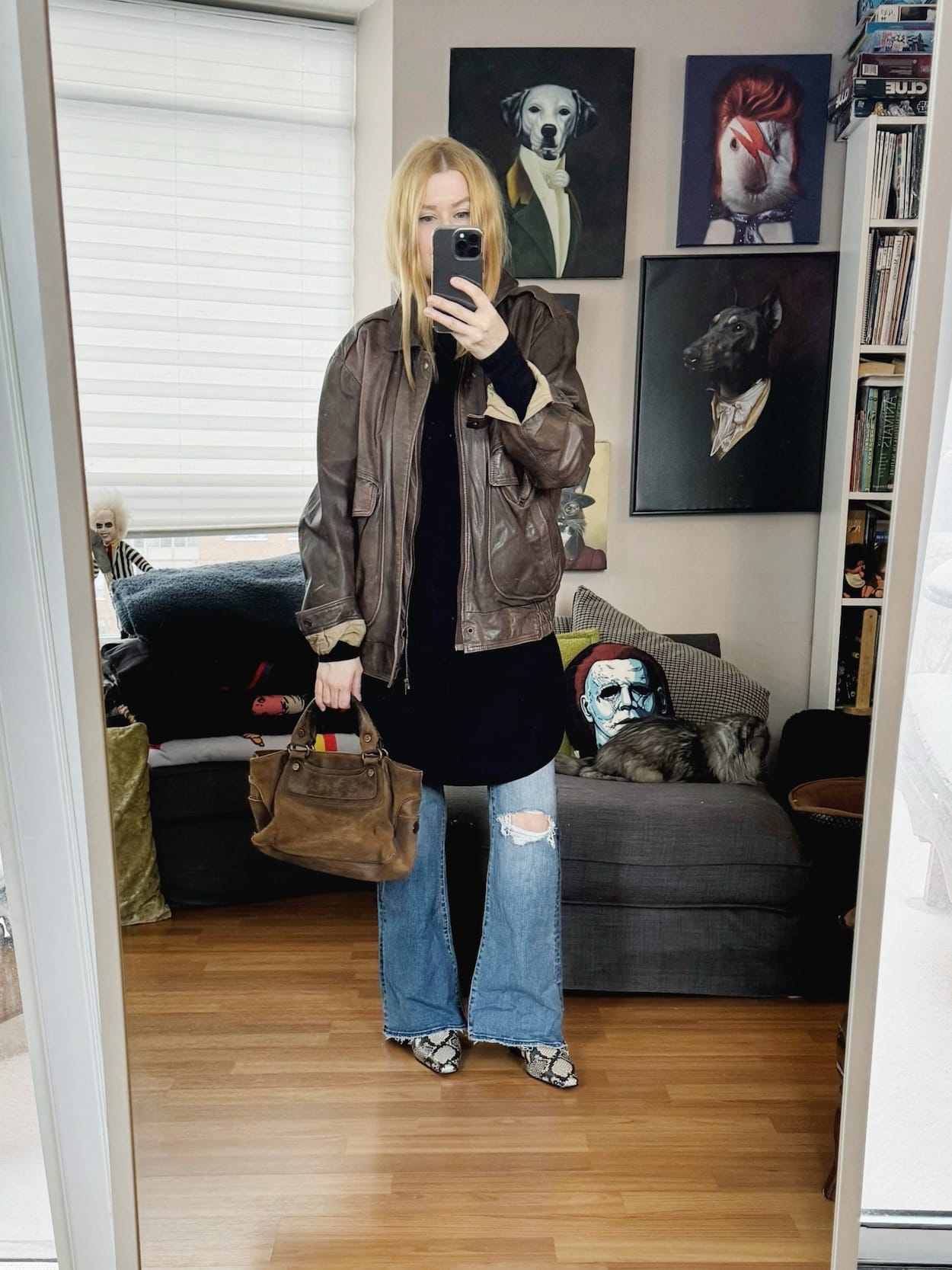 Sara of livelovesara is standing in front of a mirror taking a photo of her outfit. She is wearing a black tunic, vintage bomber jacket, flare jeans, snake print boots, and a brown suede Celine Boogie Bag.