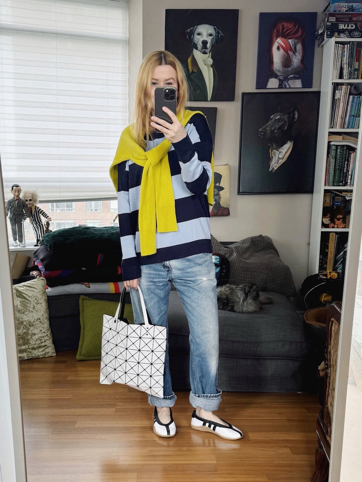 Sara of livelovesara is standing in front of a mirror taking a photo of her outfit. She is wearing a light blue and navy sweater with a bright yellow sweater tied around her shoulders, vintage Levis, Adidas ballet flats, and an Issey Miyake Bao Bao bag.