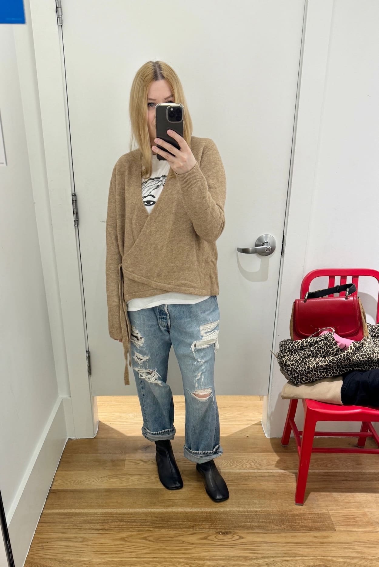 Sara of livelovesara is standing in front of a mirror taking a photo of her outfit. She is wearing a white tee under a camel wrap sweater, vintage Levis, black flats boots, an a red Fendi bag.