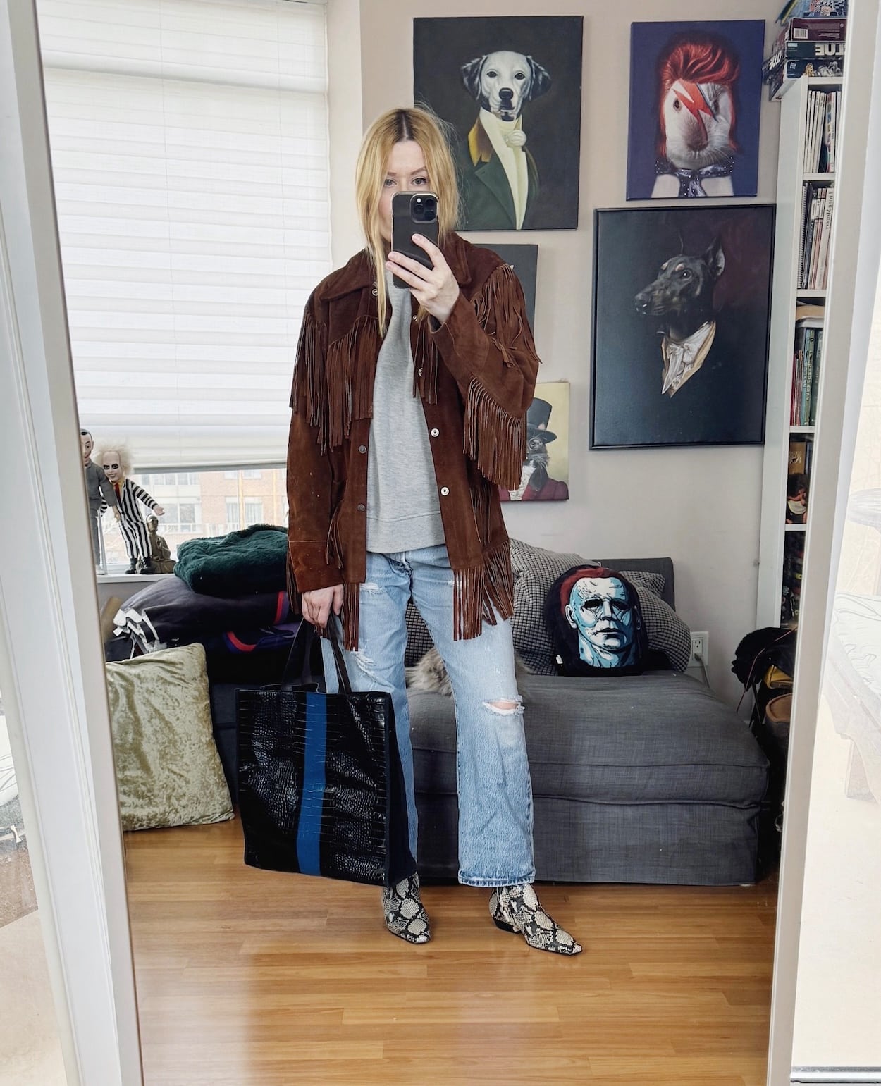 Sara of livelovesara is standing in front of a mirror taking a photo of her outfit. She is wearing a grey short sleeve sweatshirt under a vintage fringe jacket, vintage Levis, snake print boots, and a striped Dries Van Noten tote bag.
