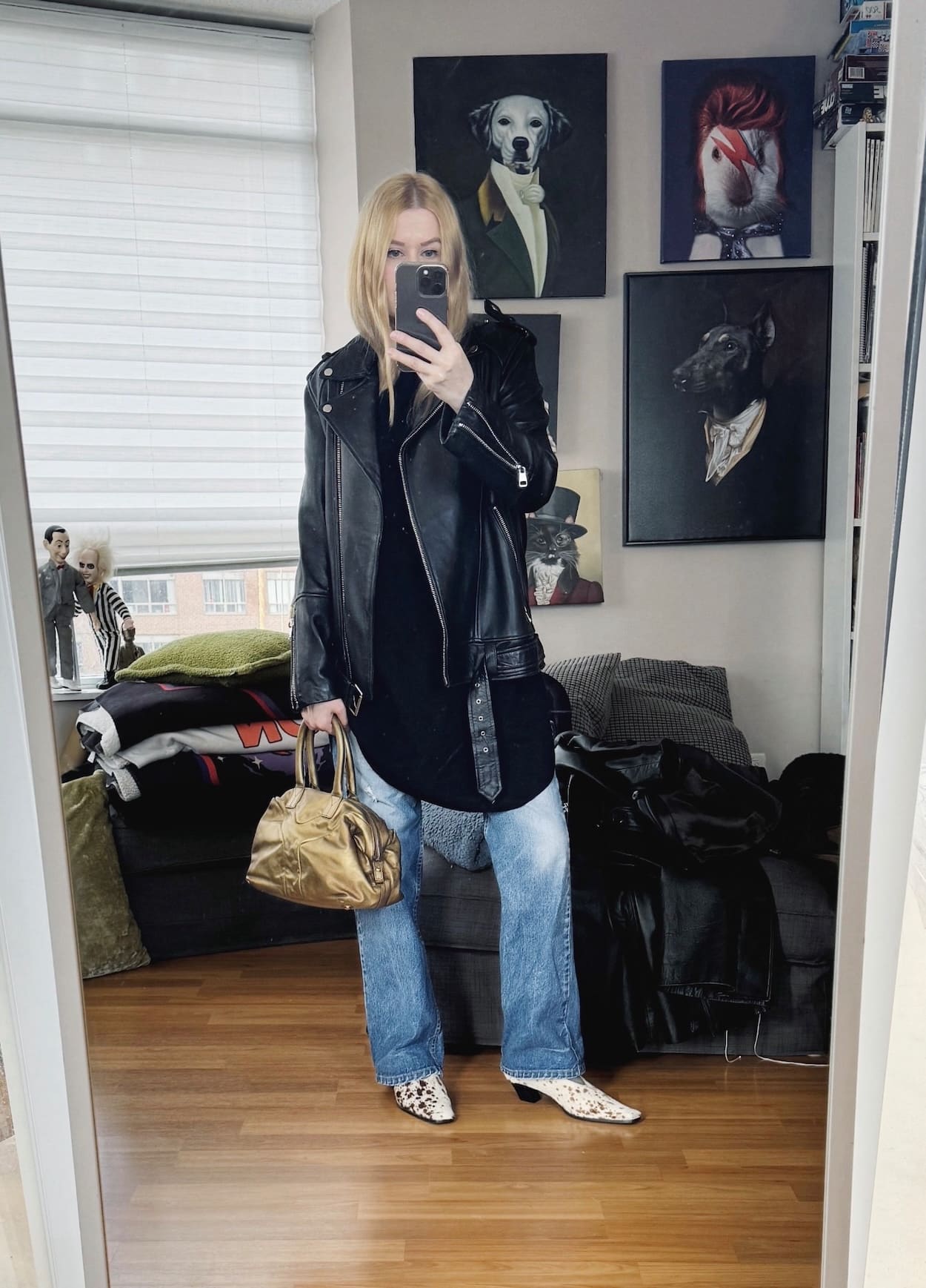 Sara of livelovesara is standing in front of a mirror taking a photo of her outfit. She is wearing a black tunic with vintage levis, a moto jacket, deer print shoes, and a gold handbag.